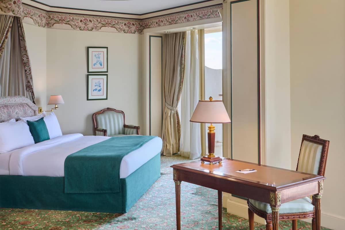 A hotel room with a bed, a table, a chair, a lamp, and framed pictures on the wall.
