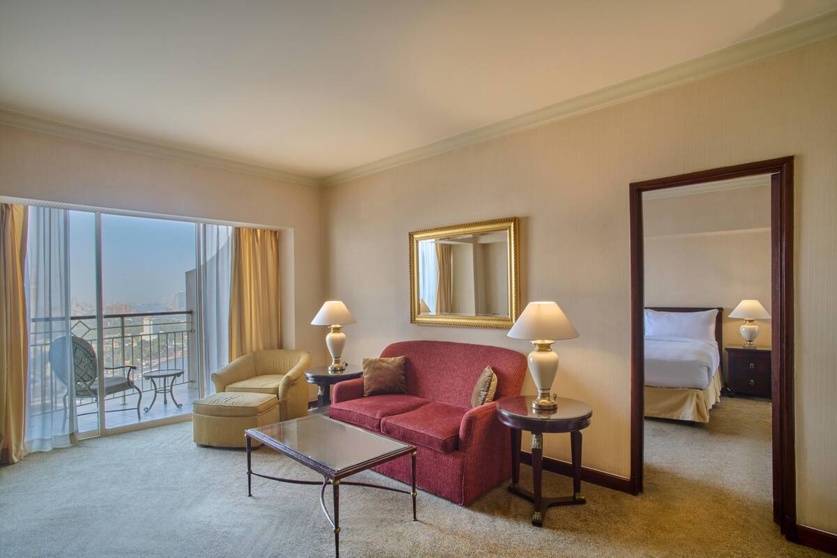 A hotel room with a red sofa, a glass table, a chair, a bed, and a large mirror.