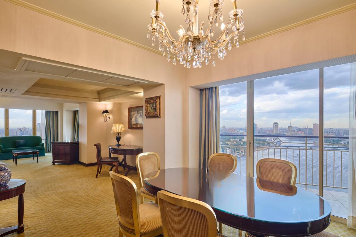 A spacious room with a large dining table, comfortable seating, a chandelier, and a view of a cityscape through large windows.
