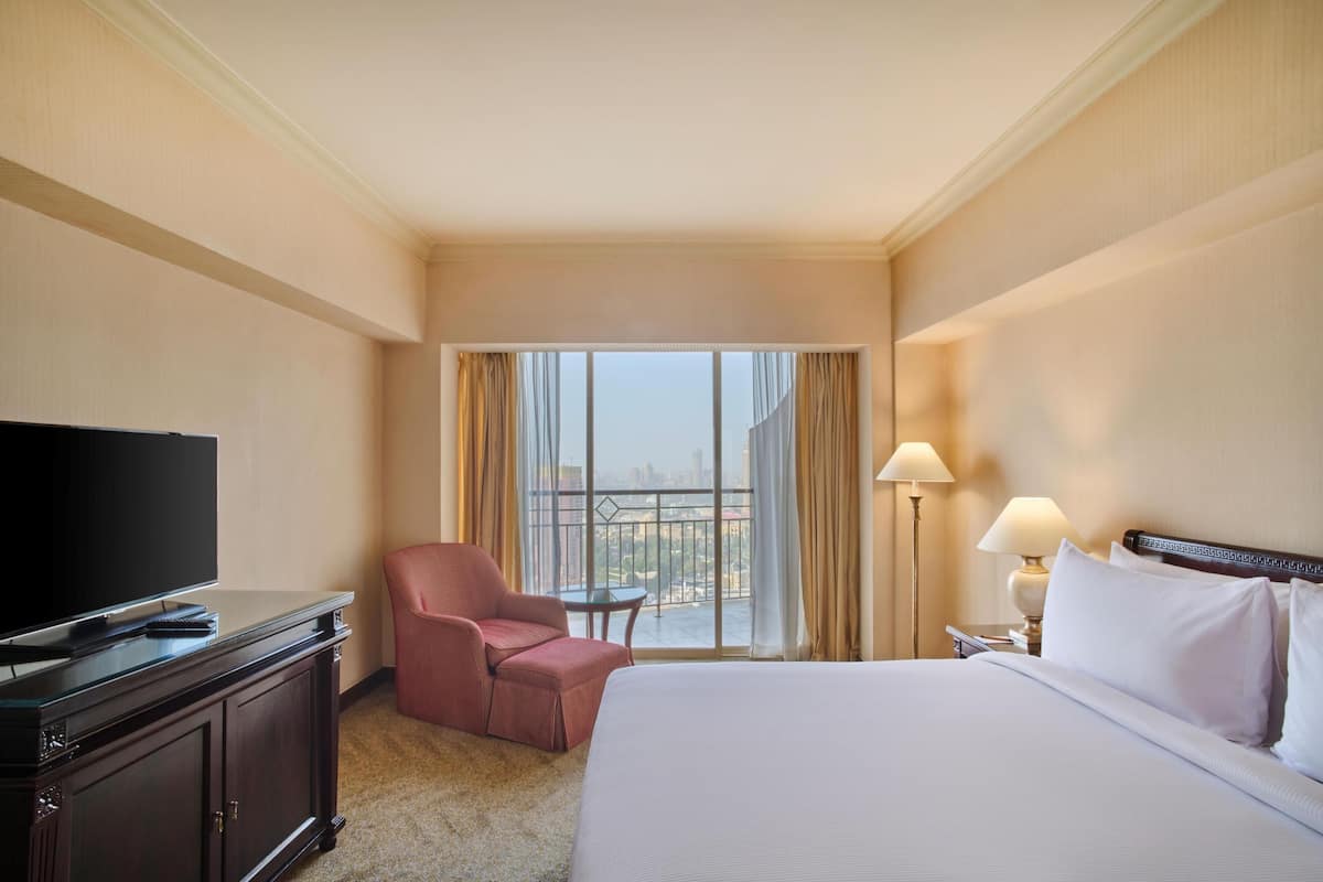 A hotel room with a bed, a television, a chair, a small table, and a view of the city through large windows.
