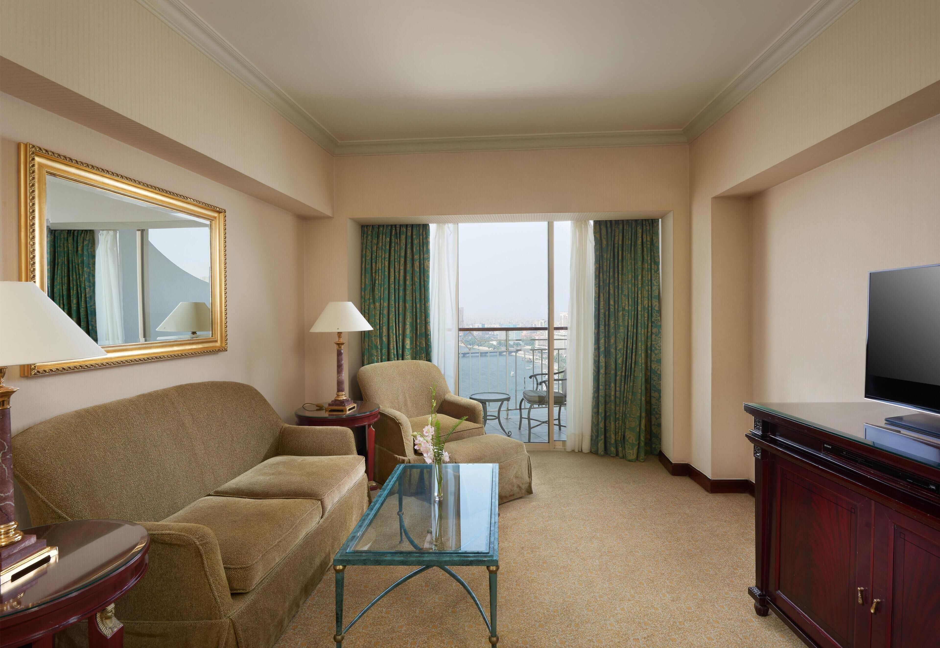 A hotel room with a sofa, armchair, glass coffee table, TV, and a balcony with curtains.