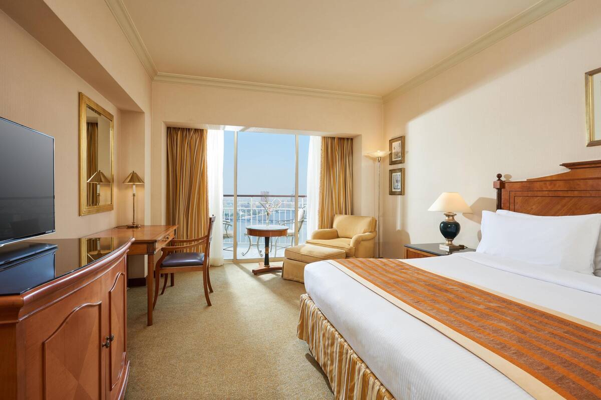 A hotel room with a bed, a TV, a desk with a chair, a small table, and a balcony with a view.