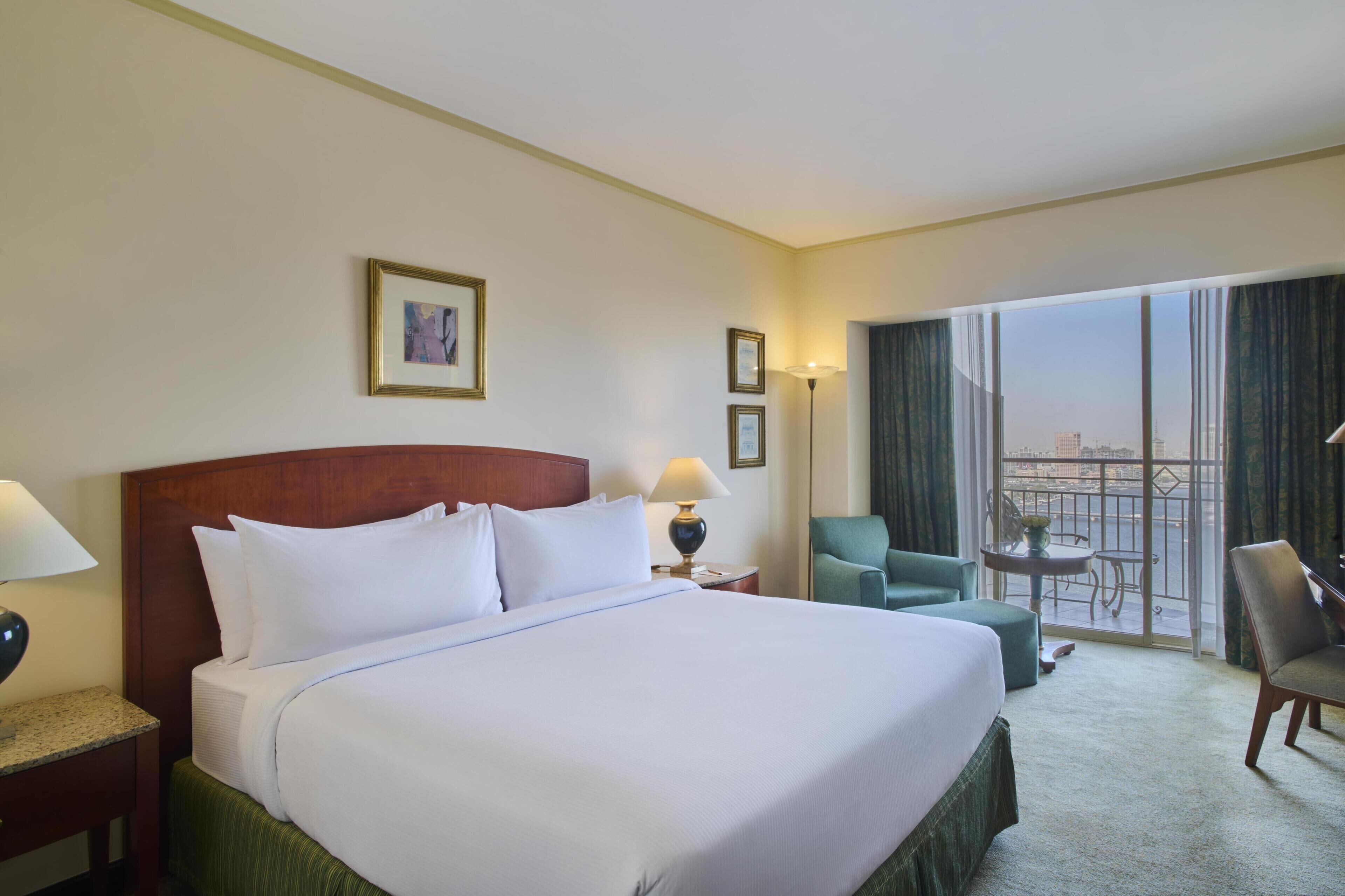 A hotel room with a large bed, a desk with a lamp, a chair, a small table, and a balcony with a view.