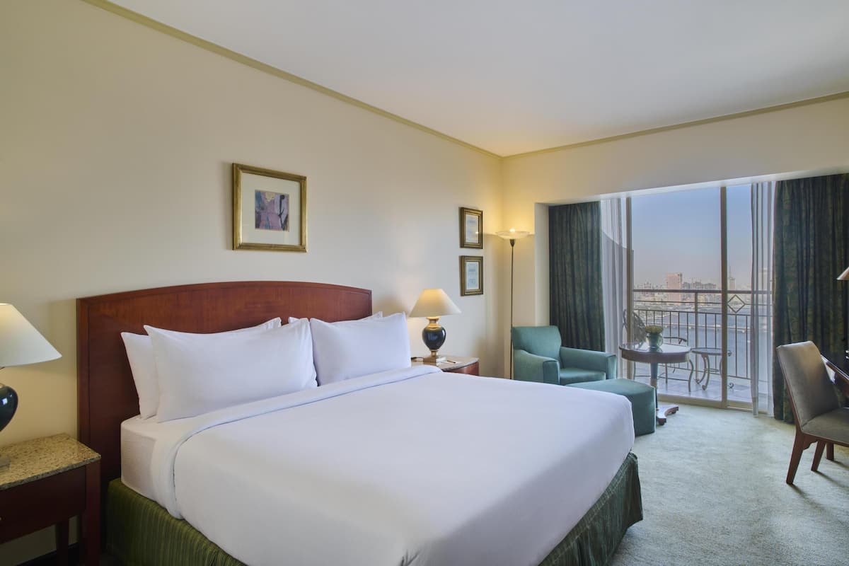 A hotel room with a large bed, a desk with a lamp, a chair, a small table, and a balcony with a view.
