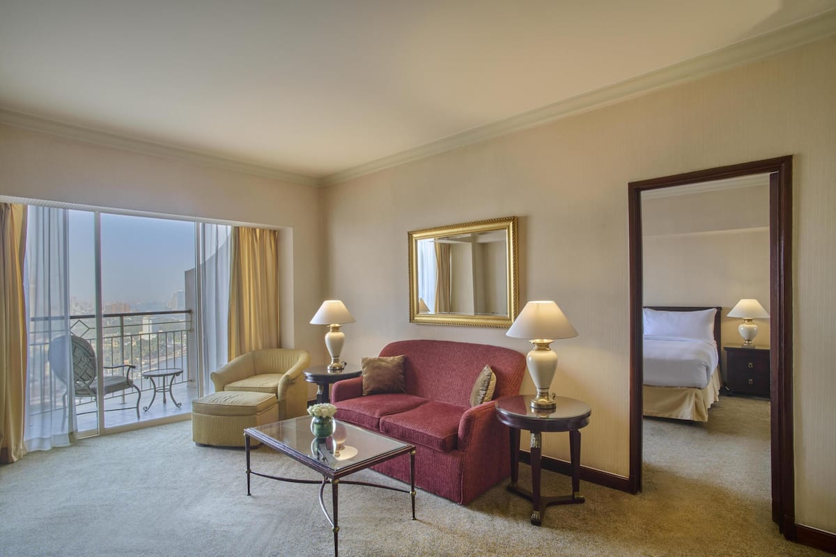 A hotel room with a red sofa, a glass coffee table, a chair, a bed, a nightstand, a lamp, a mirror, and a view of a cityscape through large windows.