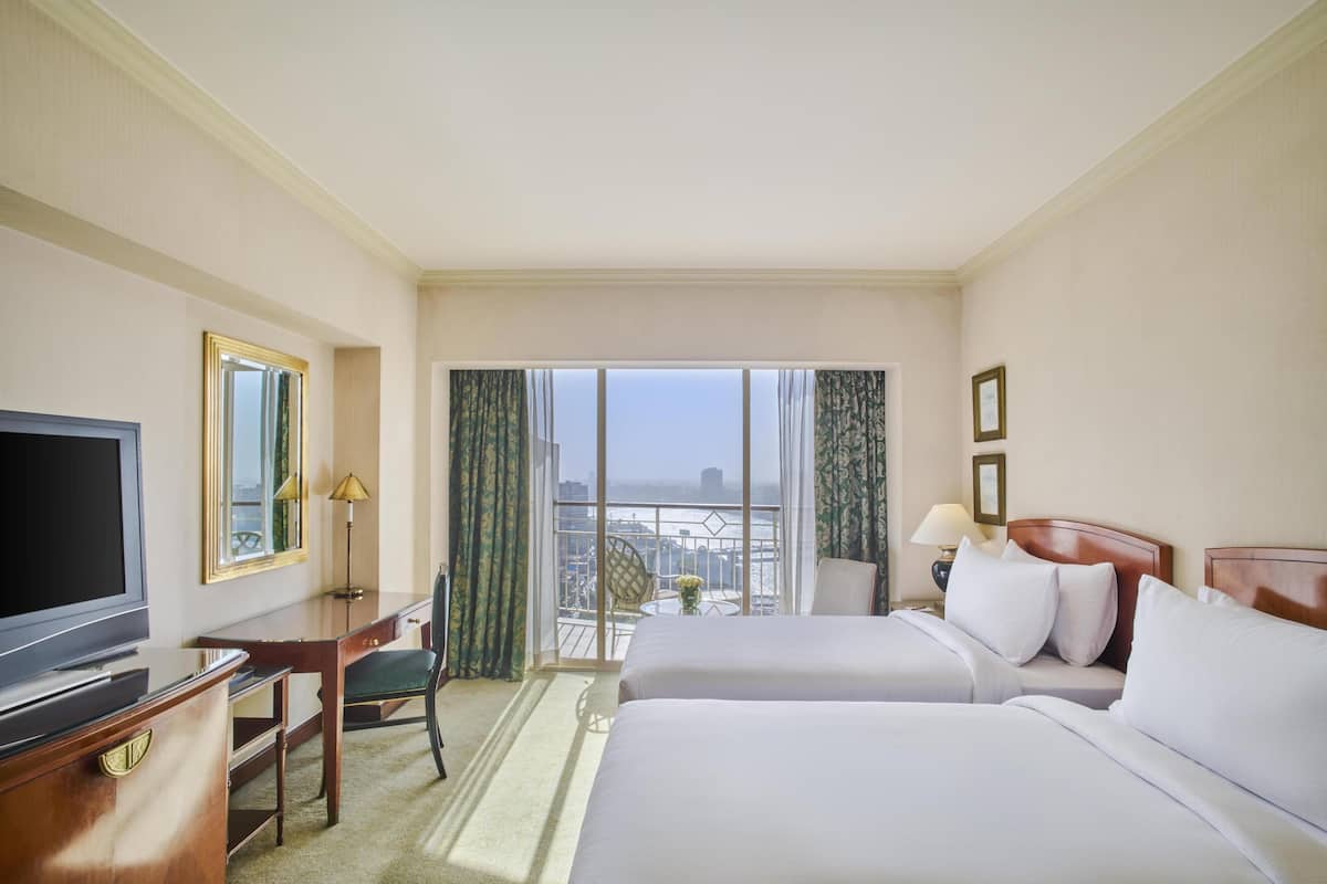 A hotel room with a TV, a desk, two beds, a chair, and a balcony with a view.