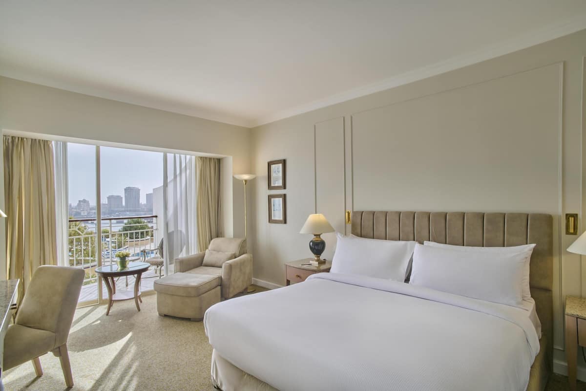 A hotel room with a large bed, a sitting area with a table and chairs, a view of the city through large windows, and a cityscape visible outside.