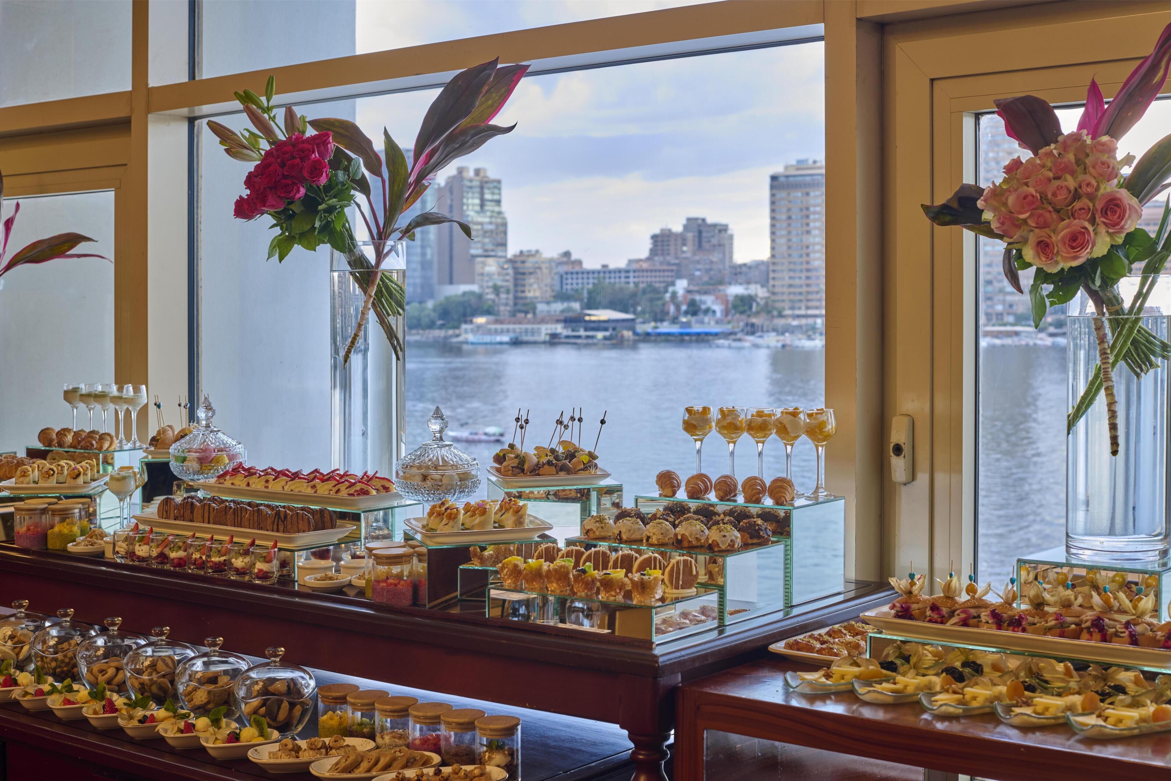 A buffet with various food items and floral decorations, with a view of a cityscape and water through large windows.