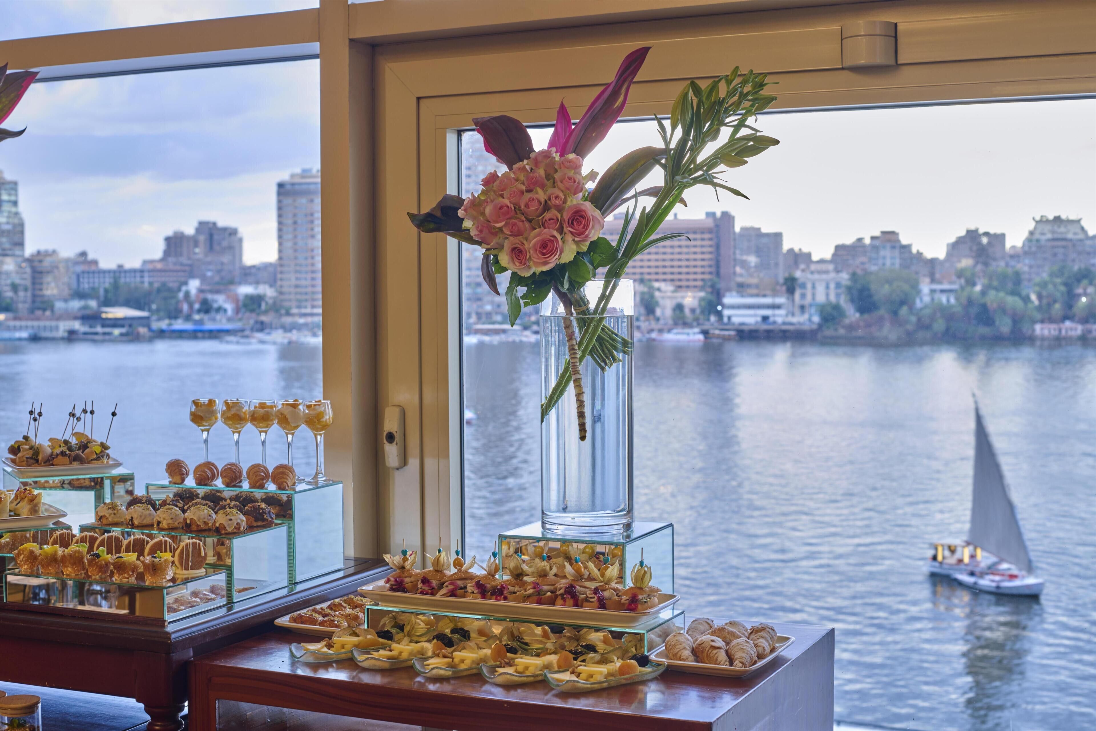 A buffet with various food items and a vase of flowers by a window overlooking a river.