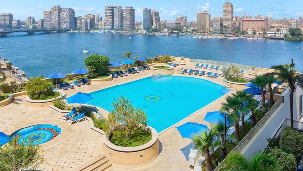 Piscina de resort con sillas de descanso y sombrillas, un horizonte urbano al fondo y un río.