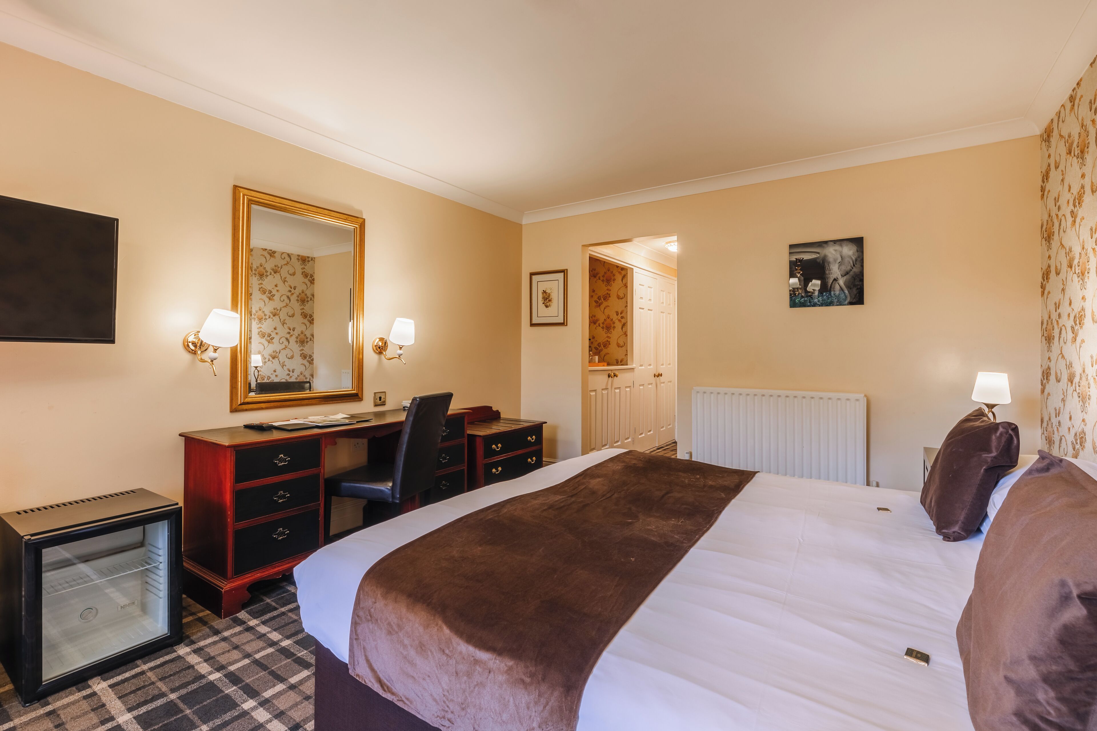 A hotel room with a bed, a desk, a chair, a dresser, a small refrigerator, and a mirror.