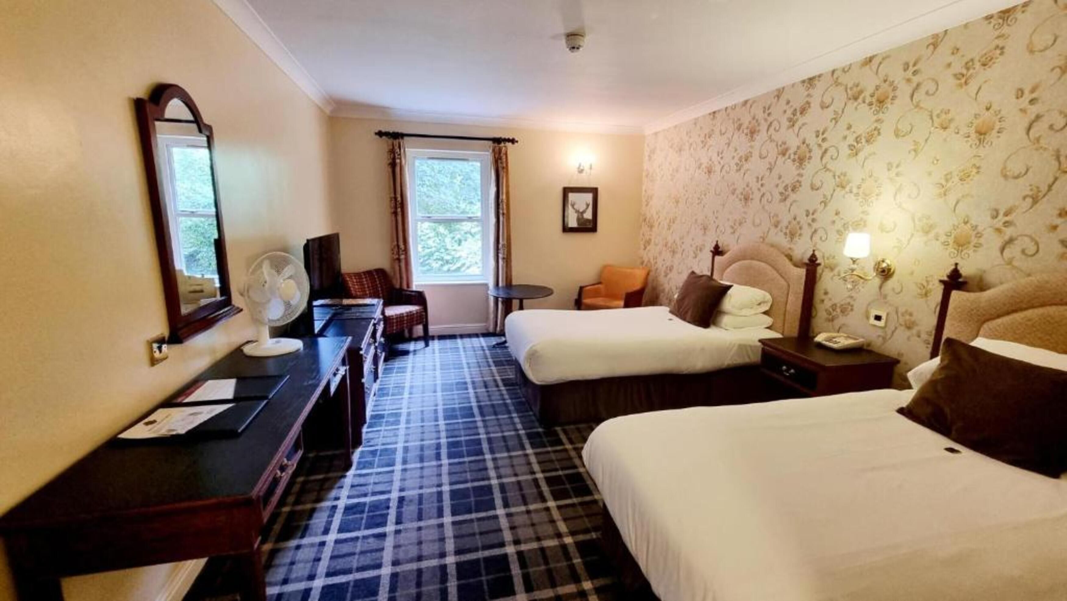 A hotel room with two beds, a desk, a chair, a fan, and a window with curtains.
