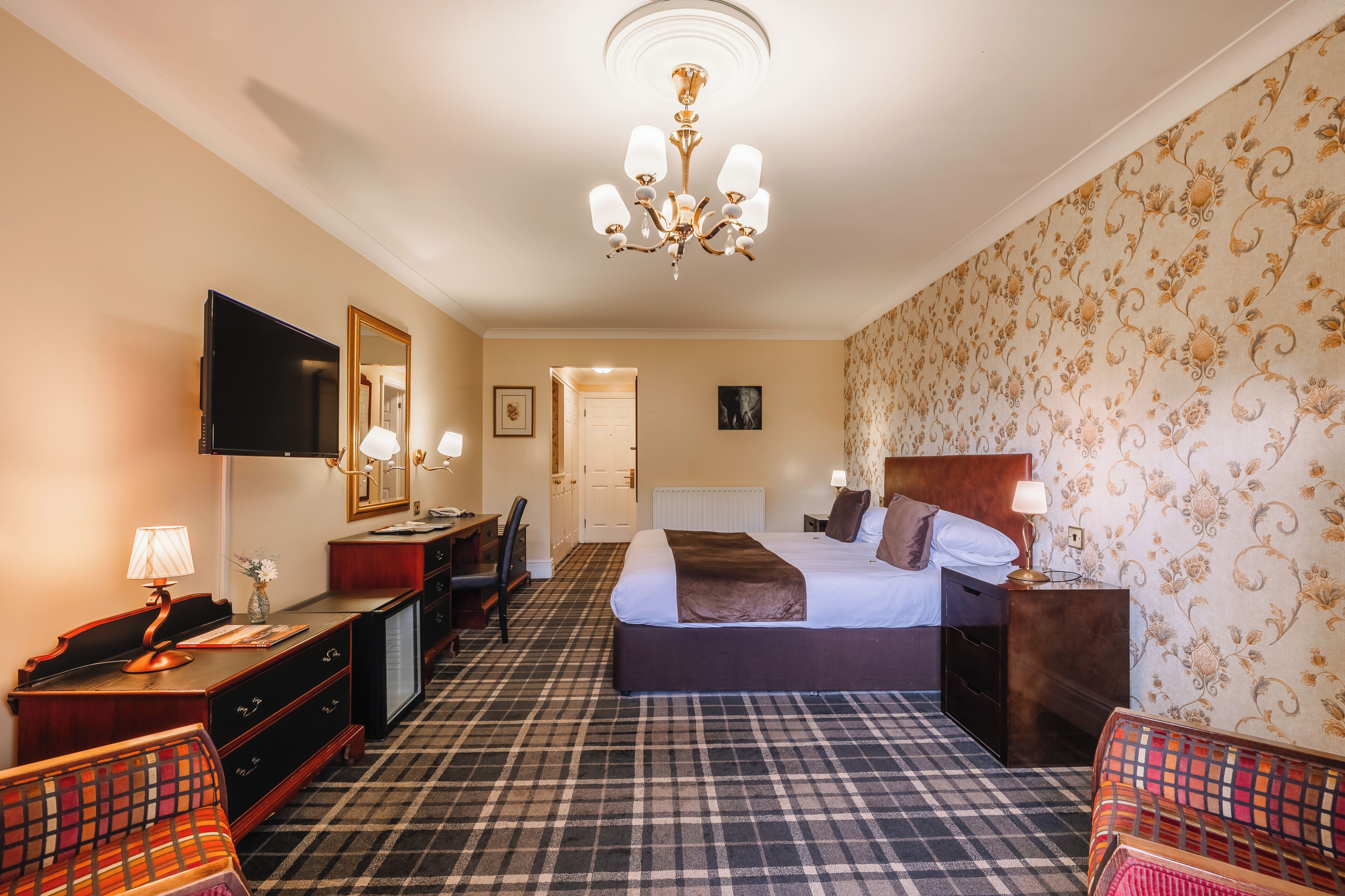 A hotel room with a checkered carpet, a bed with a headboard, a desk with a lamp, a television, and a wardrobe.