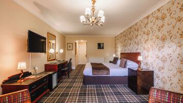 A hotel room with a checkered carpet, a bed with a headboard, a desk with a lamp, a television, and a wardrobe.
