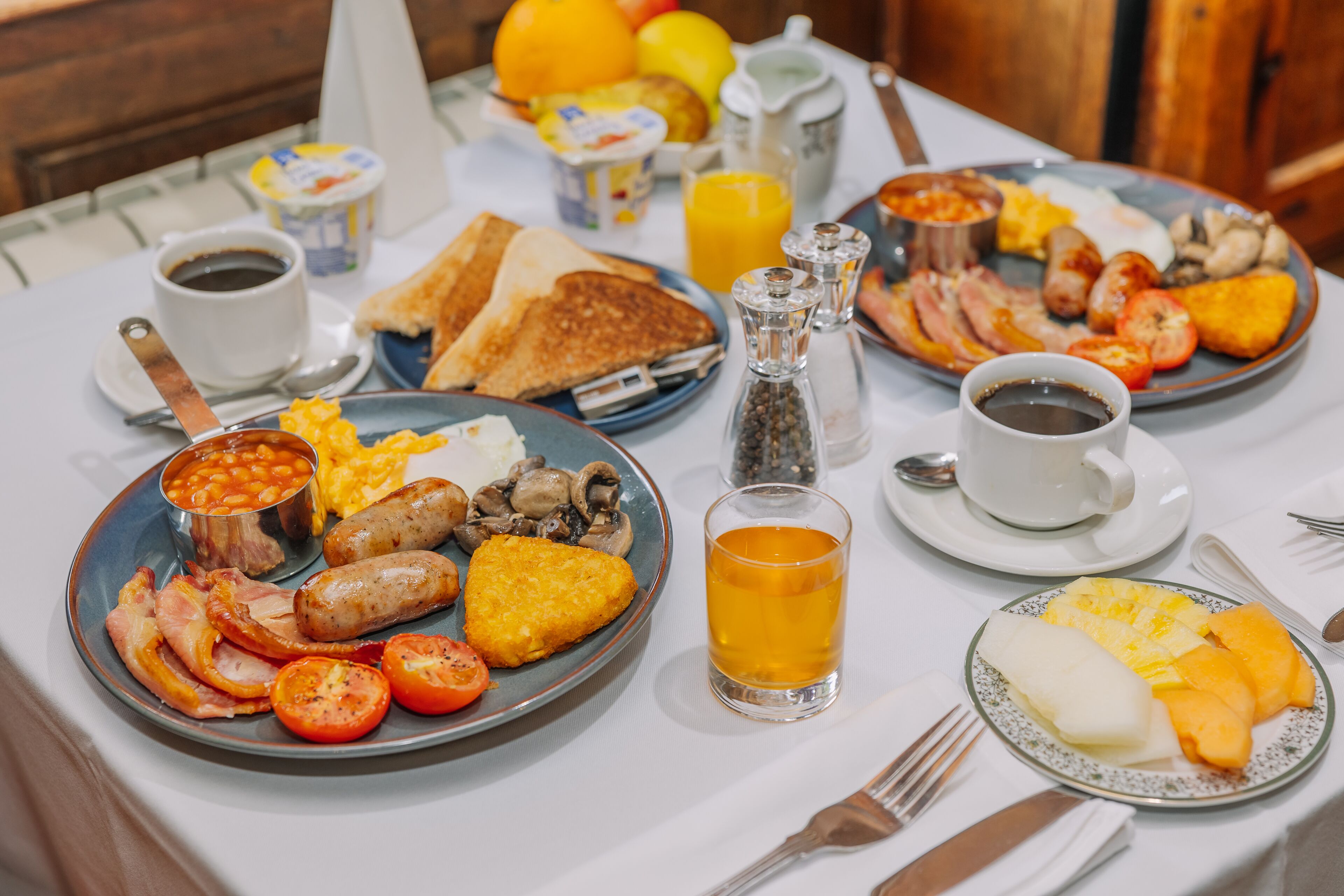 A breakfast table with plates of eggs, bacon, sausages, toast, and fruit. There are also cups of coffee and juice.