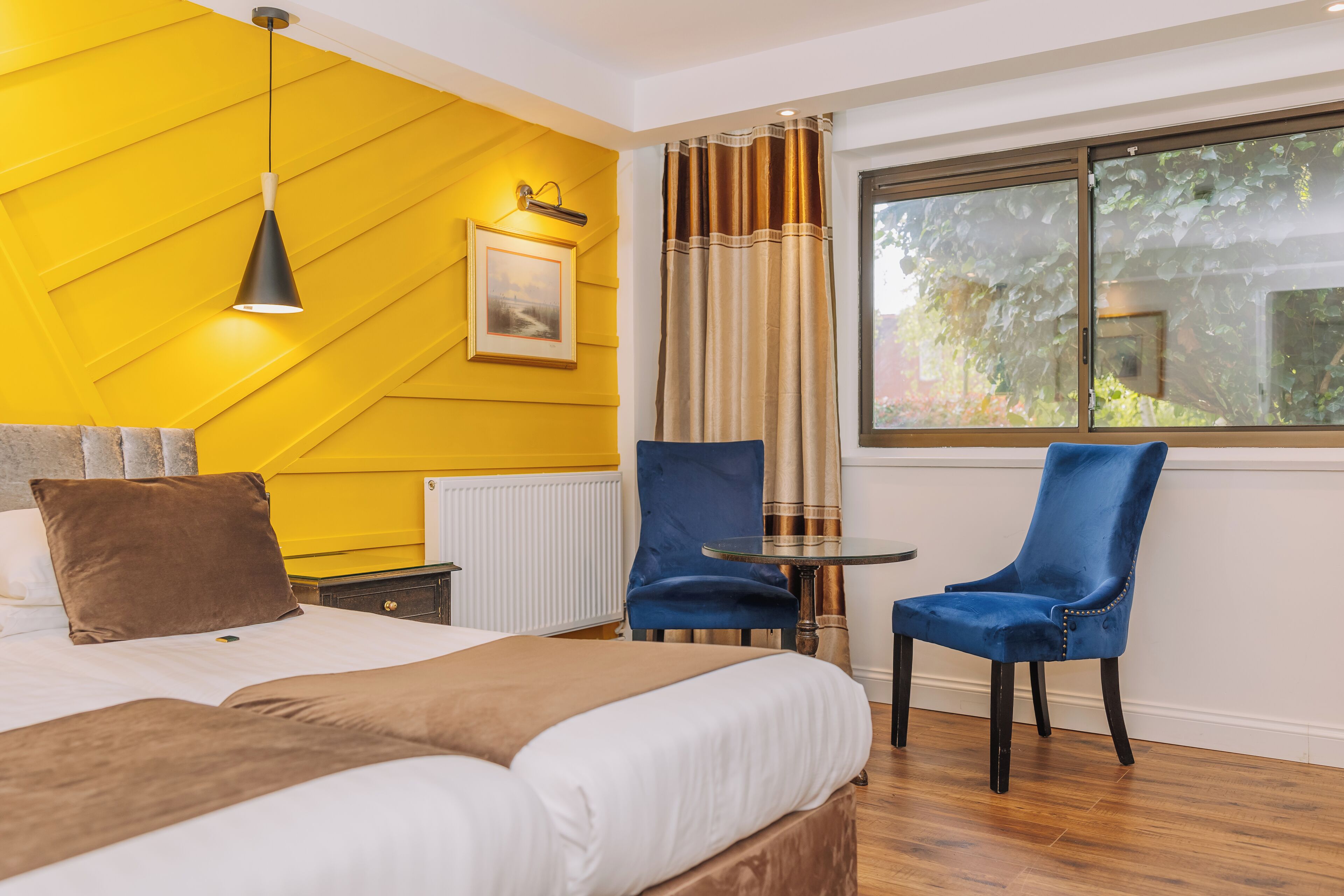 Quarto de hotel com cama, duas cadeiras, uma mesinha, janela com vista para vegetação e parede amarela.