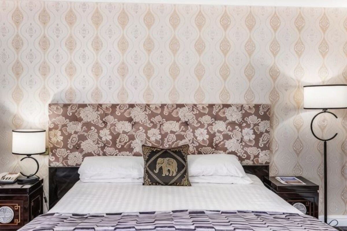 Ein Hotelzimmer mit gestreifter Tapete, einem Bett mit weißer Bettdecke, einem Kopfkissen mit Elefantenmuster, zwei Nachttischen mit Lampen und einem Beistelltisch mit einer Uhr.