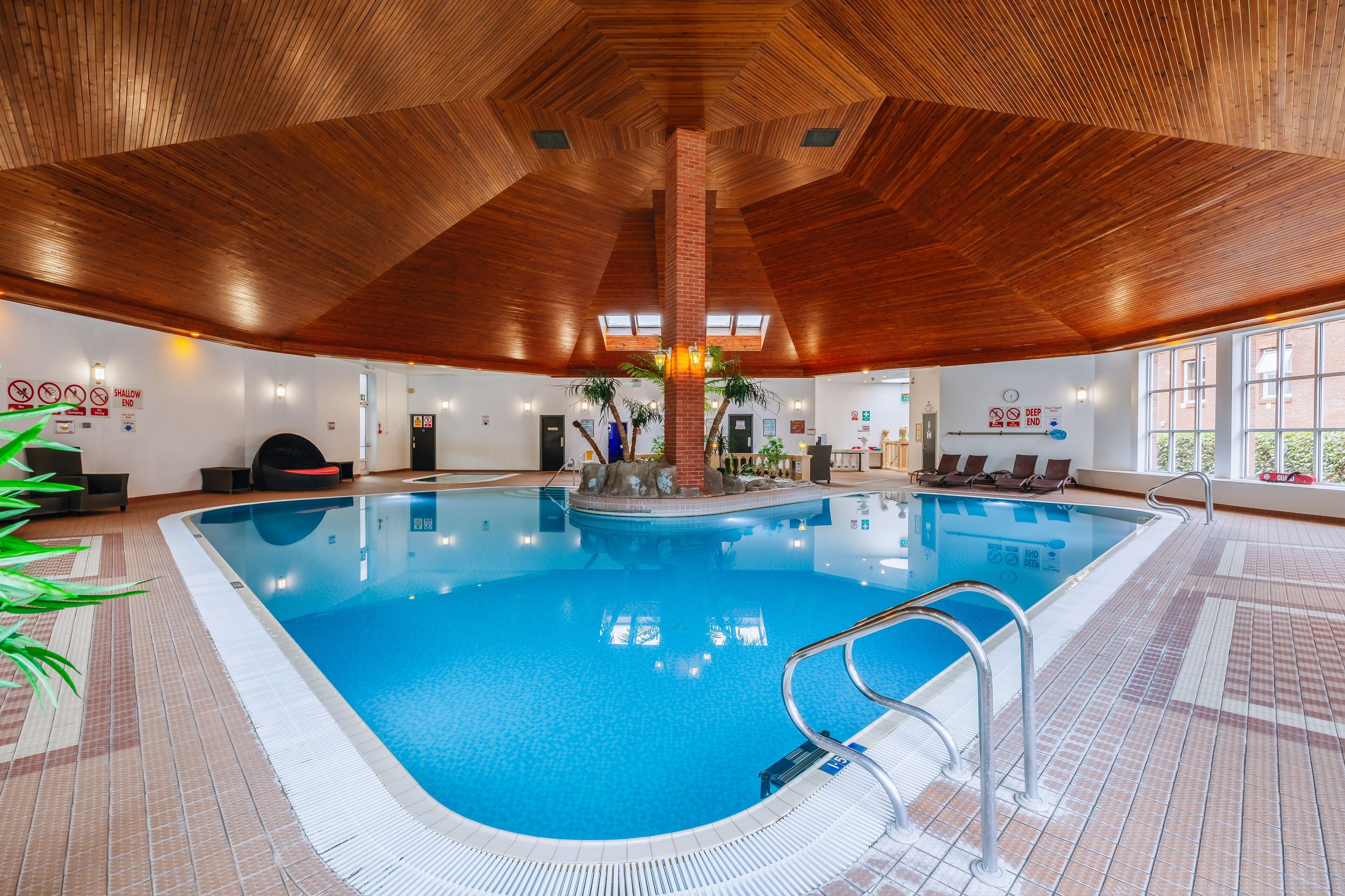 Una piscina interna con soffitto in legno e pavimento piastrellato, circondata da poltrone relax e piante in vaso.