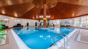 Una piscina interna con soffitto in legno e pavimento piastrellato, circondata da poltrone relax e piante in vaso.