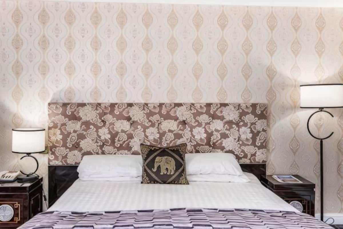 A hotel room with a patterned wallpaper, a bed with a white bedspread, a pillow with an elephant design, two bedside tables with lamps, and a nightstand with a clock.