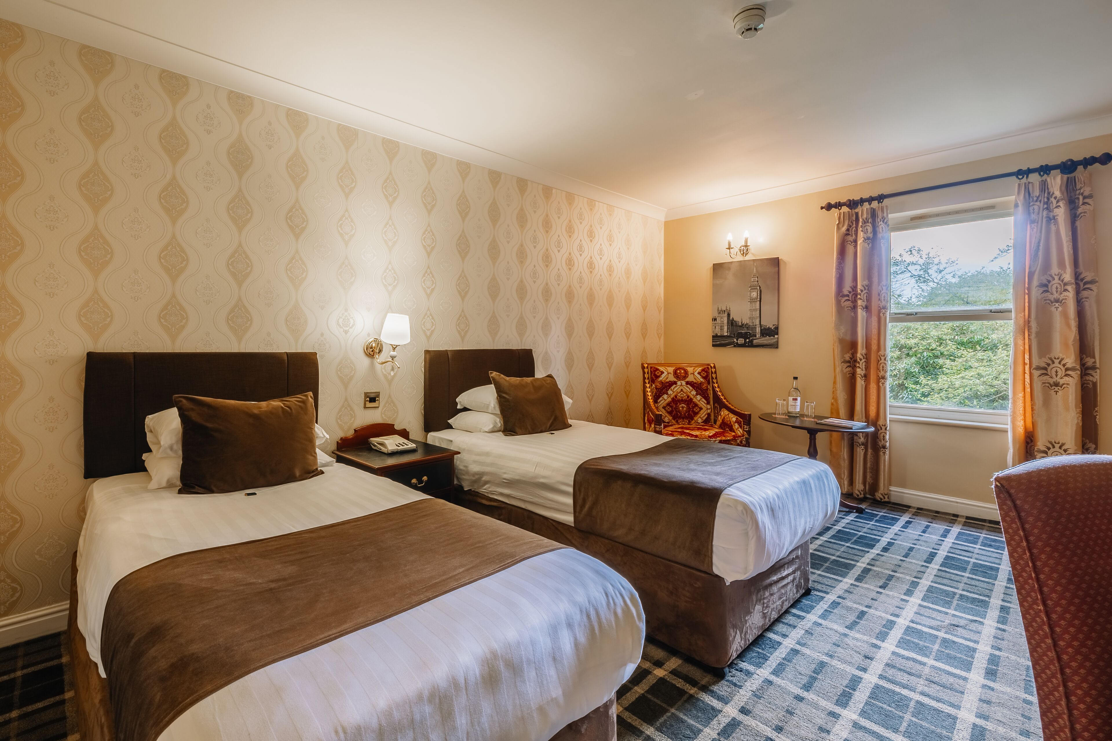 A hotel room with two beds, a small table with a lamp, a chair, and a window with curtains.