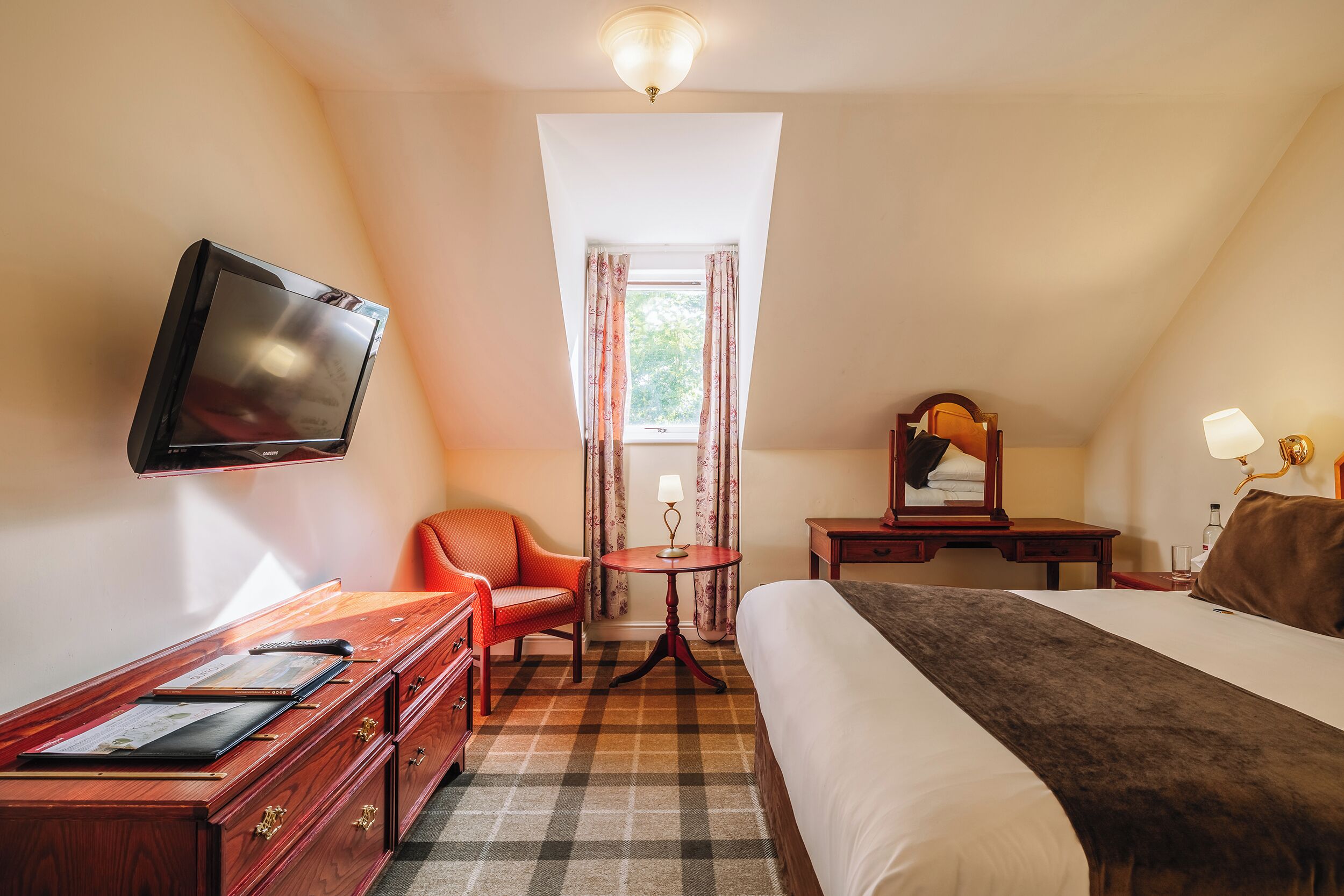 A hotel room with a bed, a desk with a TV, a chair, a small table with a lamp, and a window with curtains.
