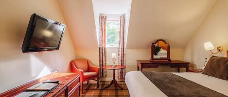 A hotel room with a bed, a desk with a TV, a chair, a small table with a lamp, and a window with curtains.