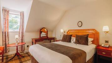 Una habitación de hotel con una cama grande, un cabecero de madera, una mesita de noche con lámpara, una pequeña mesa con lámpara, una silla y una ventana con cortinas.