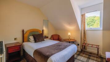 A hotel room with a bed, a chair, a small table with a lamp, a window with curtains, and a coat rack.