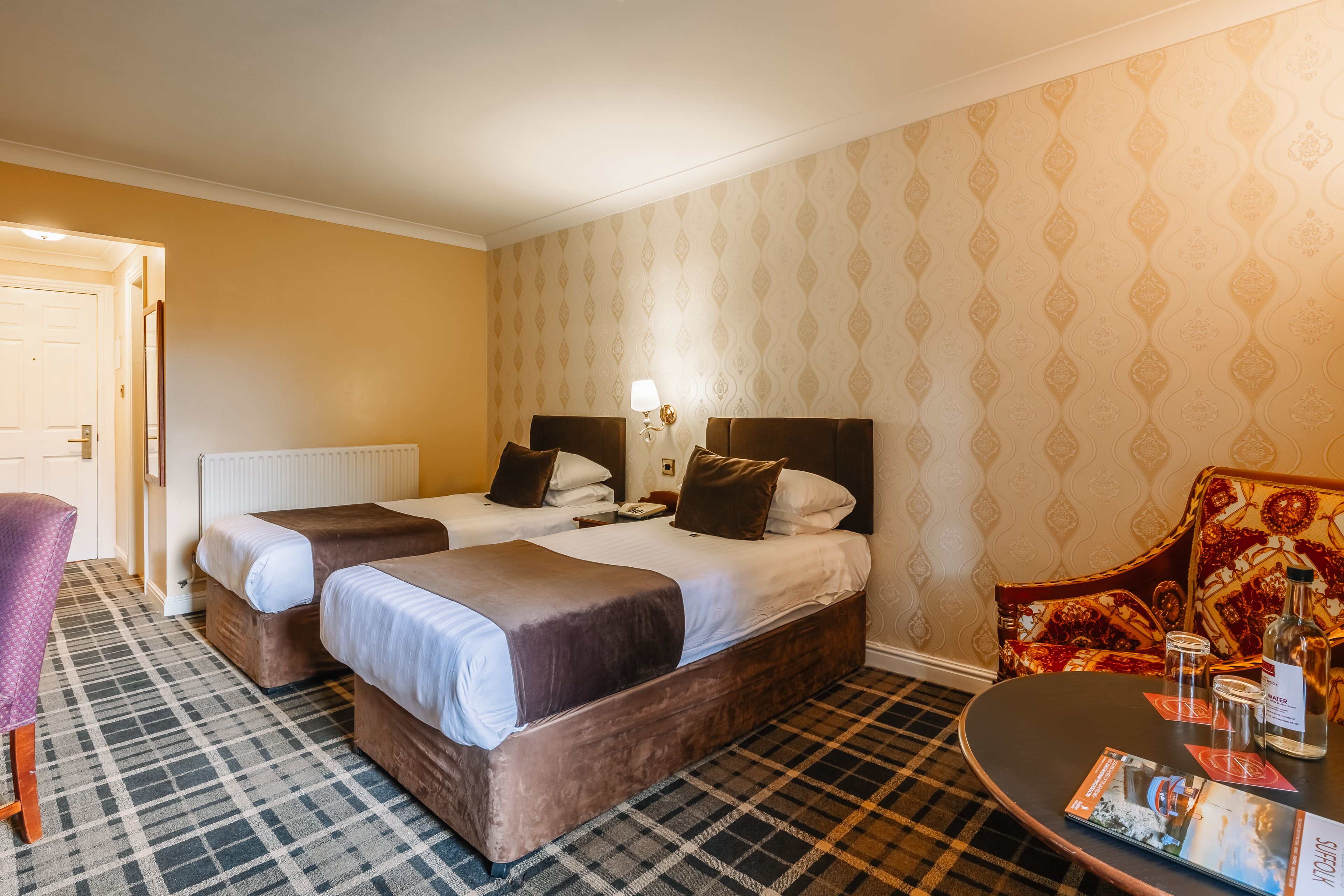 A hotel room with two beds, a checkered floor, a patterned wall, and a table with a bottle and glasses.