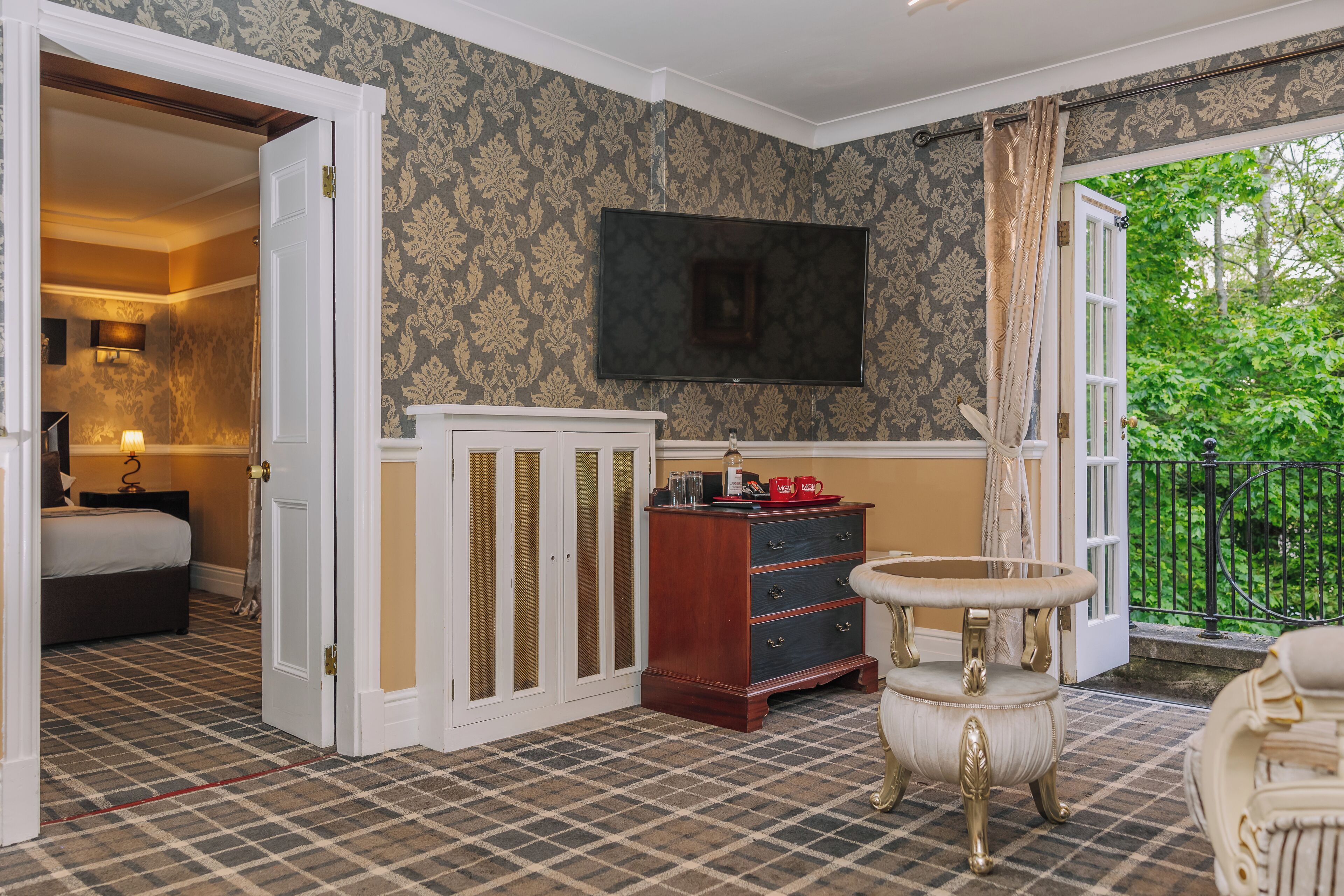 A hotel room with a checkered floor, a bed, a dresser, a television, and a balcony with a view of greenery.