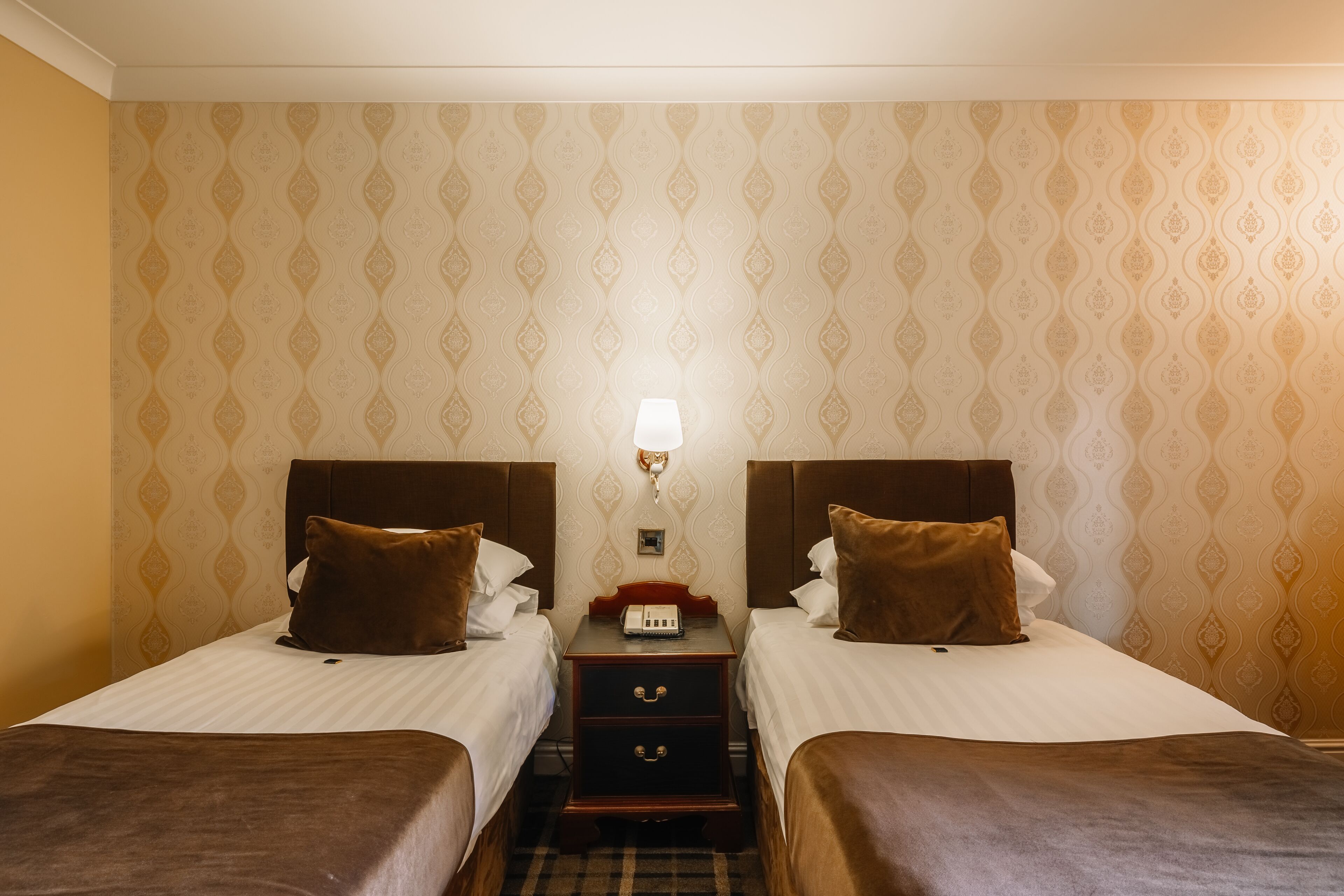 Een hotelkamer met twee bedden, een nachtkastje met een lamp en behang met een patroon.