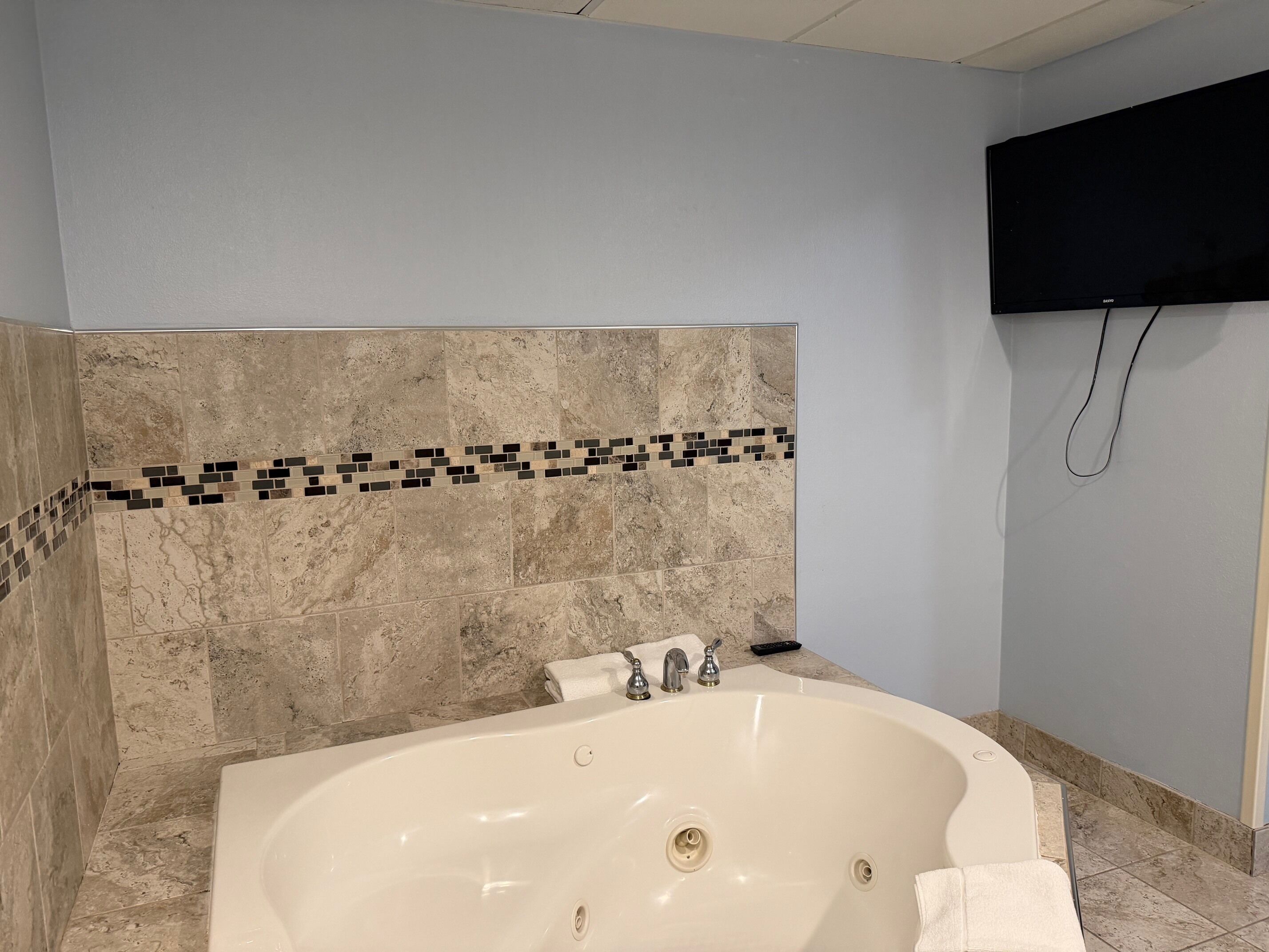 A bathroom with a bathtub, a tiled wall, a flat-screen TV mounted above the tub, and a towel rack.