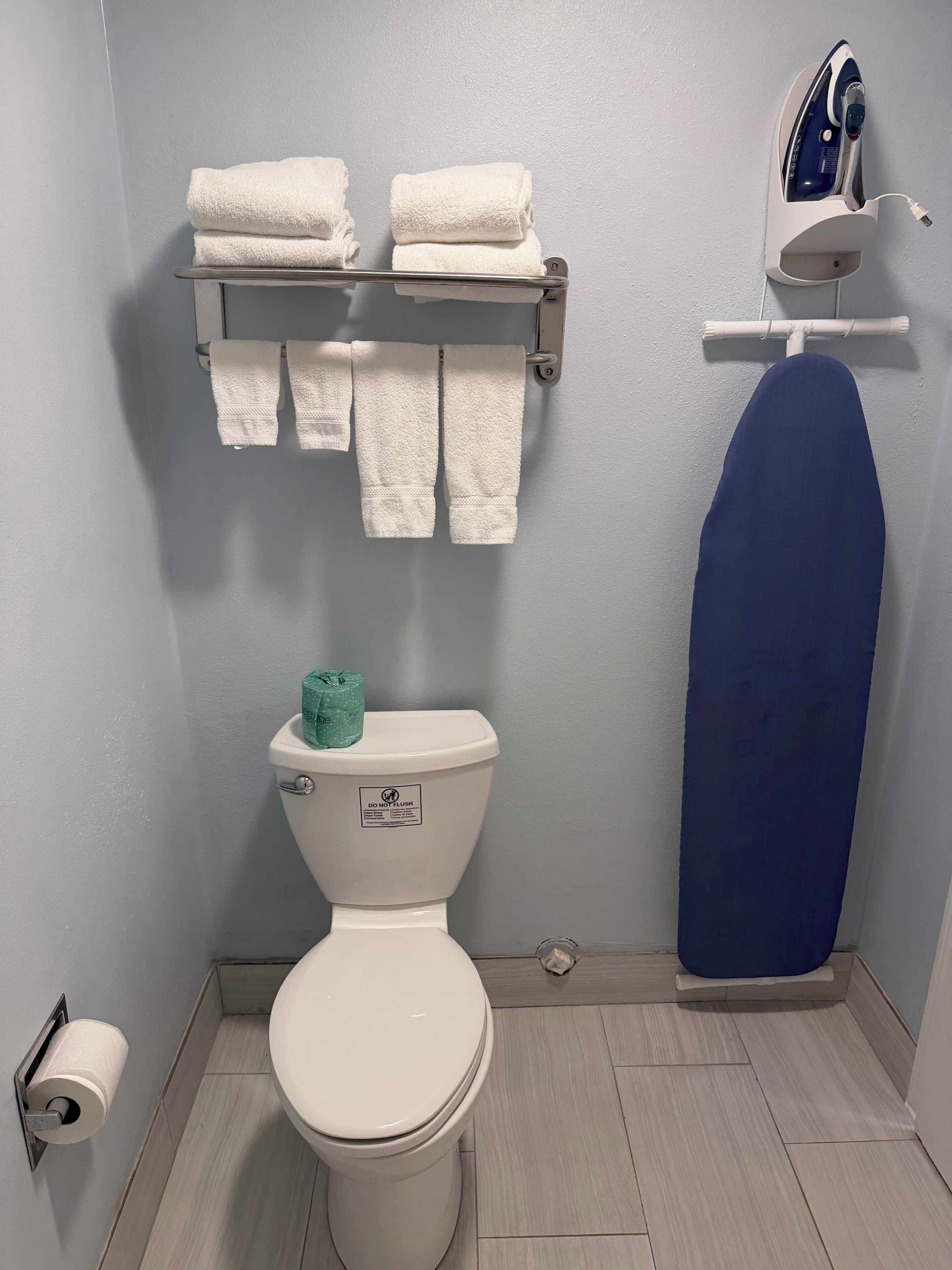 Un baño con inodoro, un perchero con toallas dobladas, un producto de limpieza verde y una tabla de planchar.
