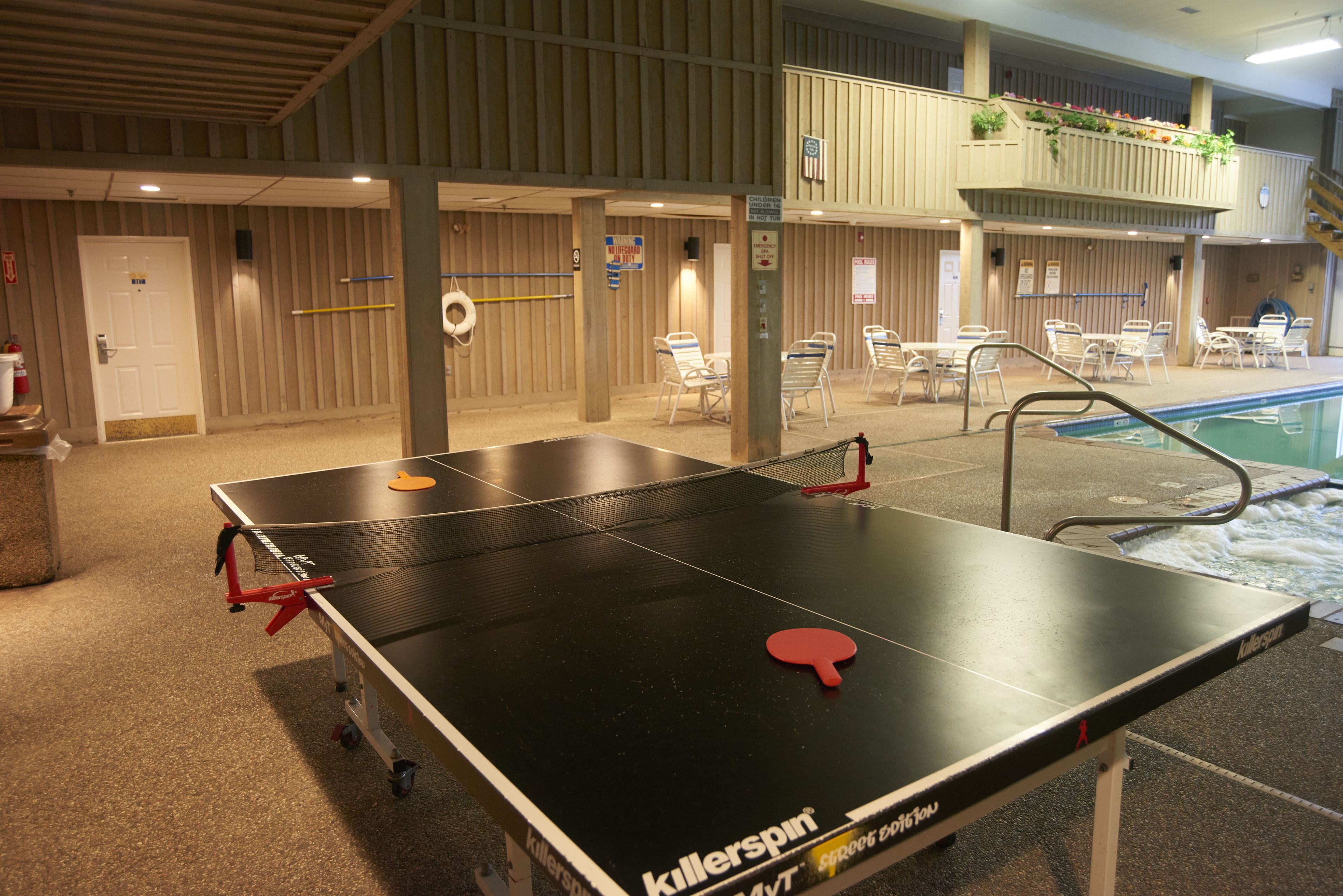A ping pong table with a red paddle, located in a room with a pool and lounge chairs.