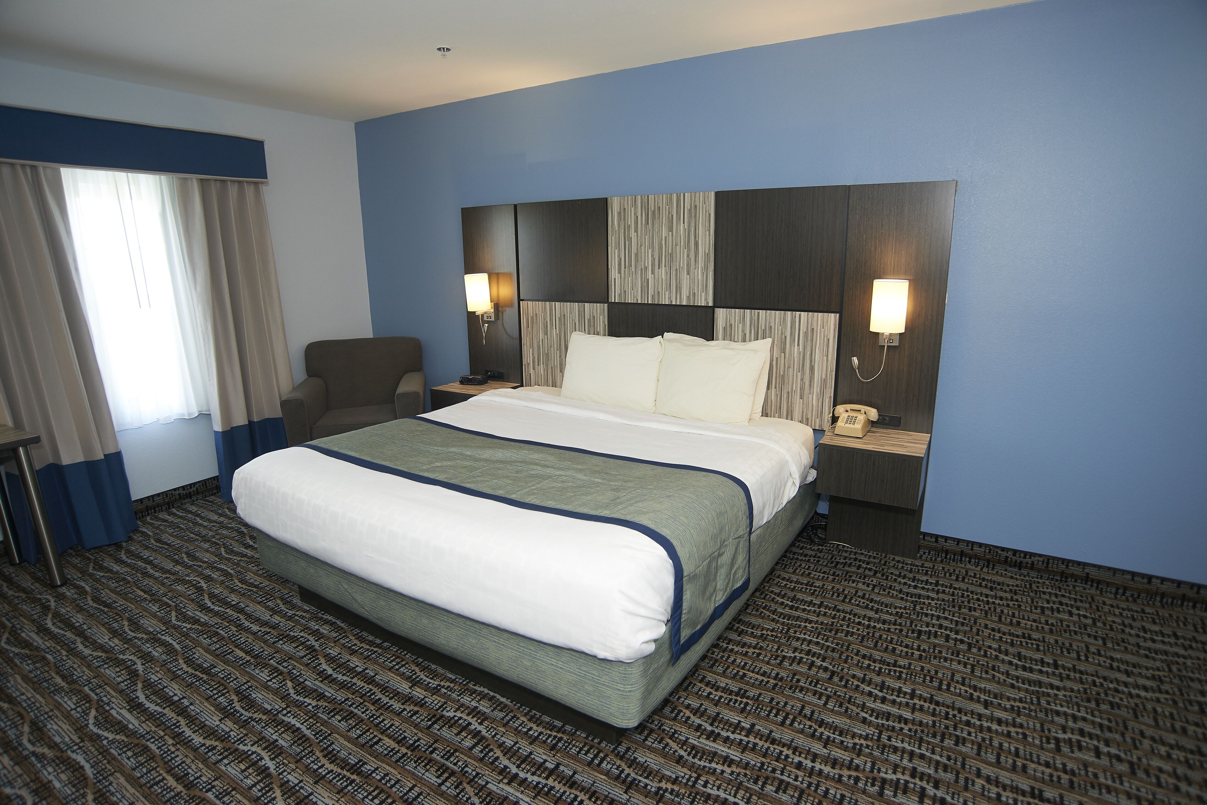Habitación de hotel con una cama grande, dos mesitas de noche con lámparas, una silla y una alfombra estampada.