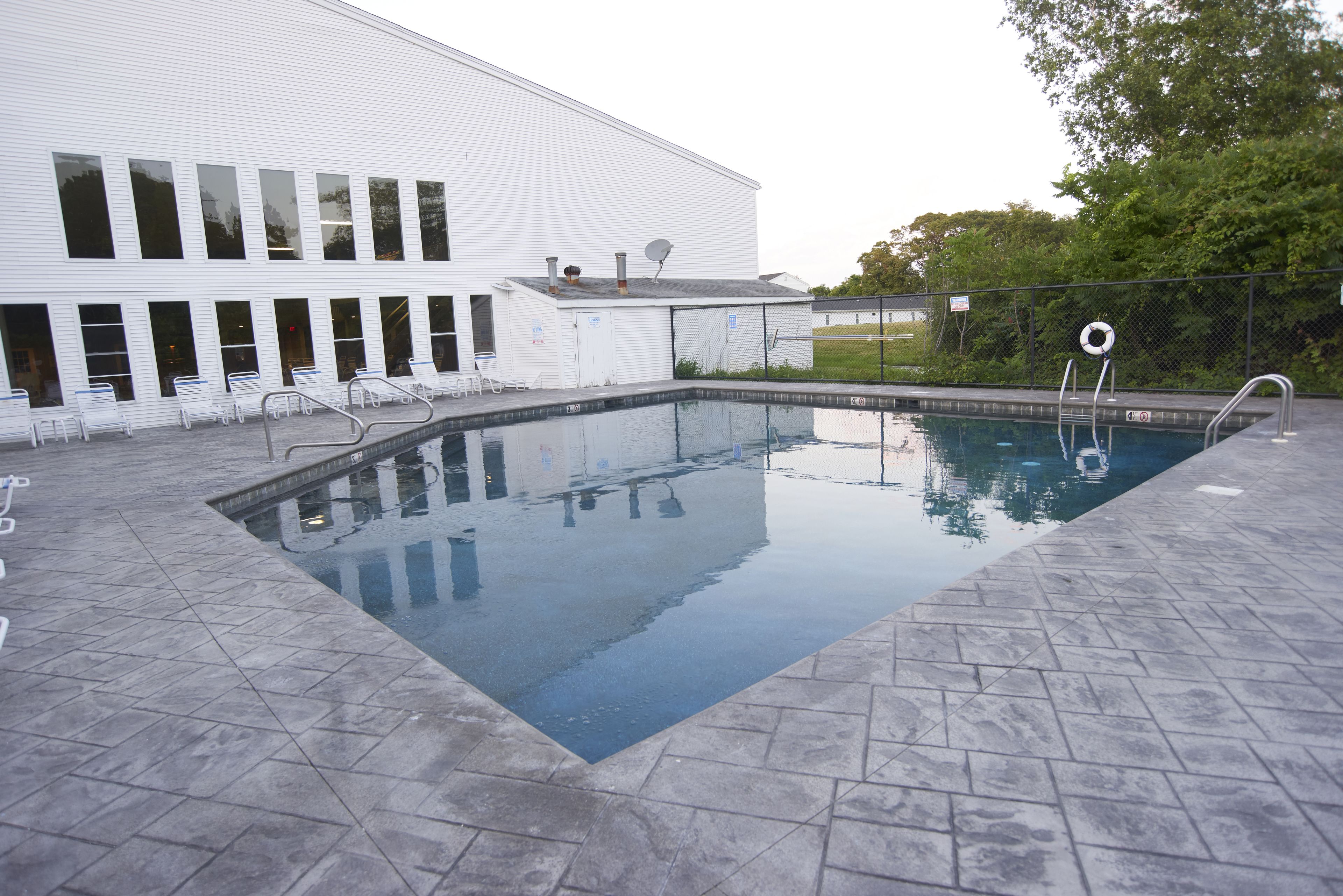 A rectangular swimming pool with a tiled deck, a white building with multiple windows and doors, and a fence with a lifebuoy.
