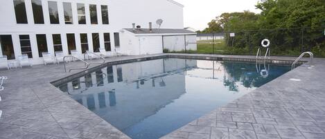 A rectangular swimming pool with a tiled deck, a white building with multiple windows and doors, and a fence with a lifebuoy.