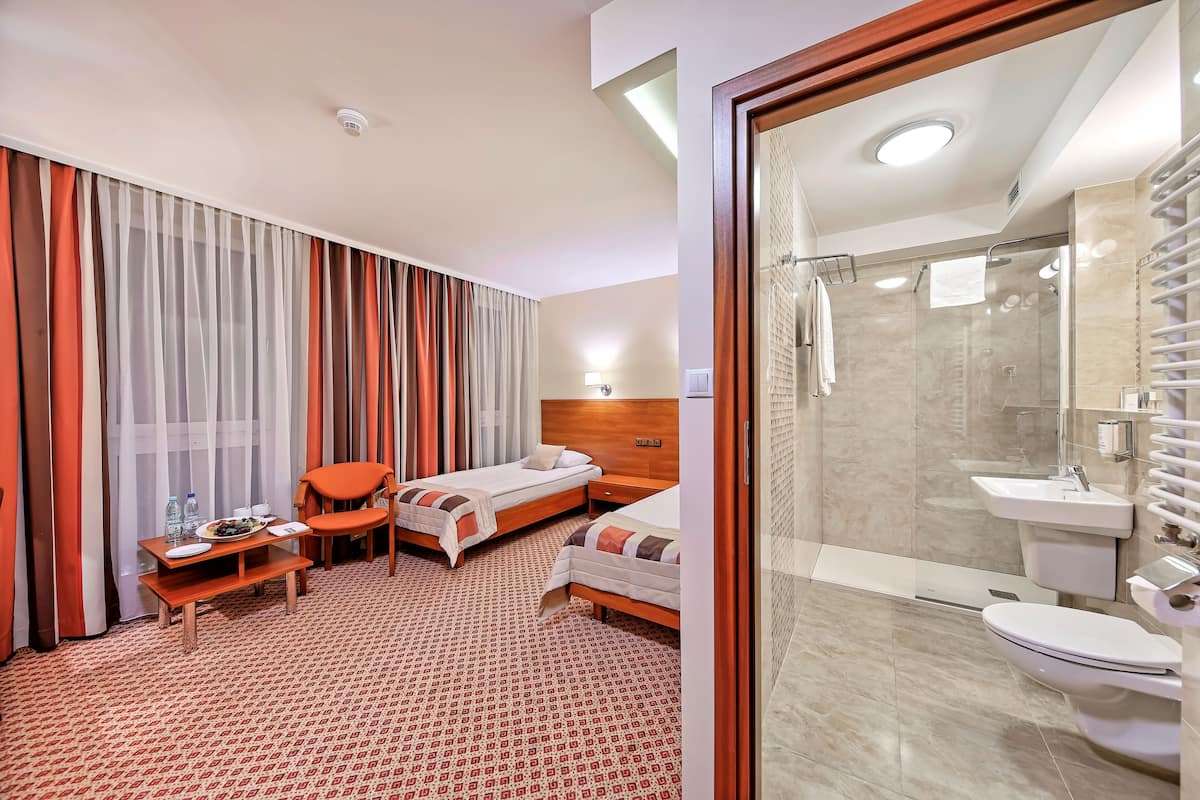 A hotel room with two beds, a small table with a chair, a shower area, and a bathroom with a sink and toilet.