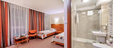 A hotel room with two beds, a small table with a chair, a shower area, and a bathroom with a sink and toilet.