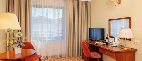 A hotel room with a desk, two chairs, a television, a lamp, a vase with flowers, and a window with curtains.