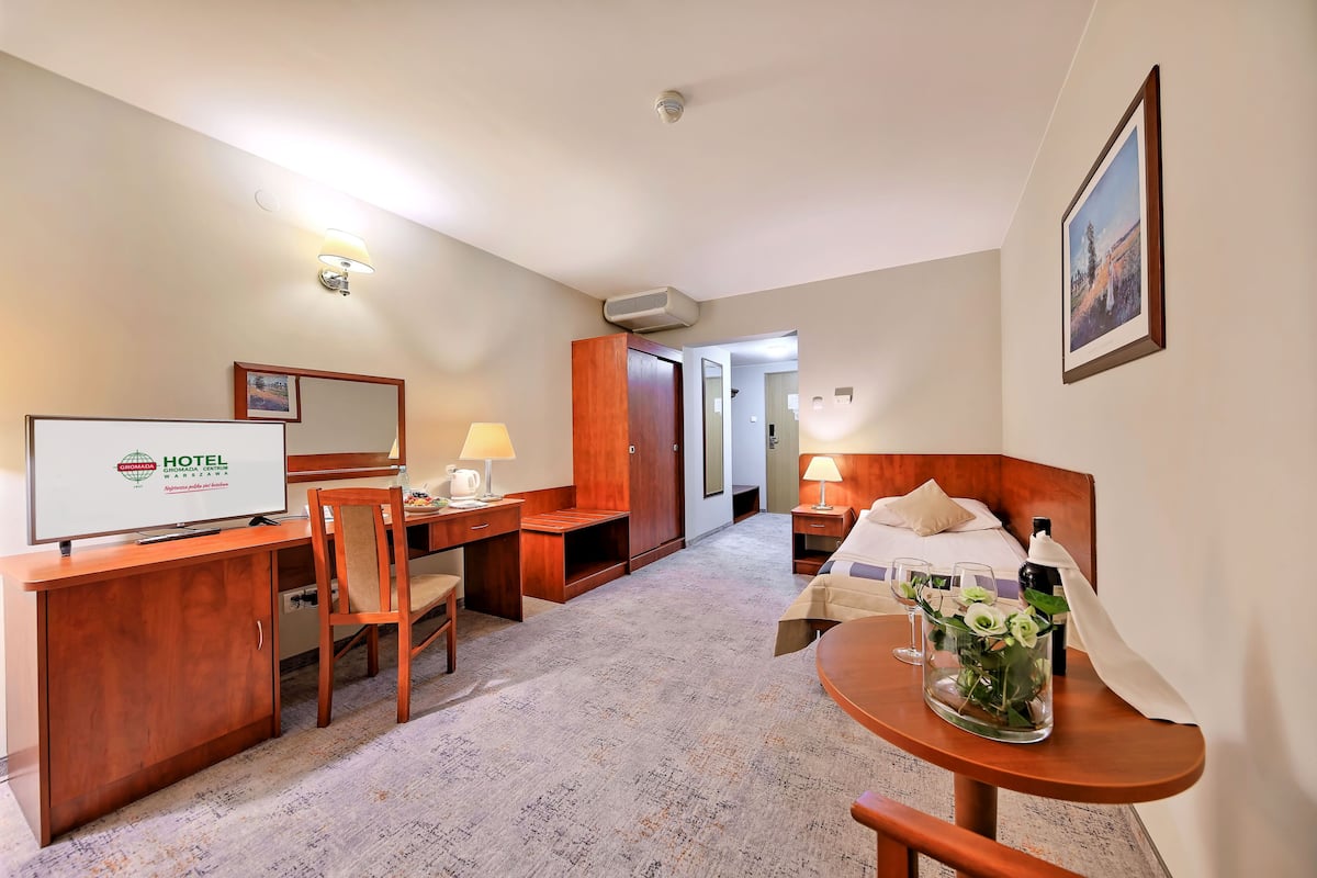 A hotel room with a desk, chair, bed, bedside table with a vase of flowers, and a television.