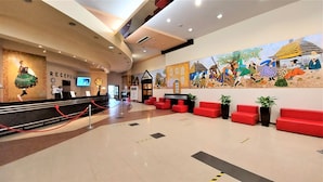Vestíbulo de hotel con mostrador de recepción, zona de estar en rojo y un mural que representa una escena tradicional.