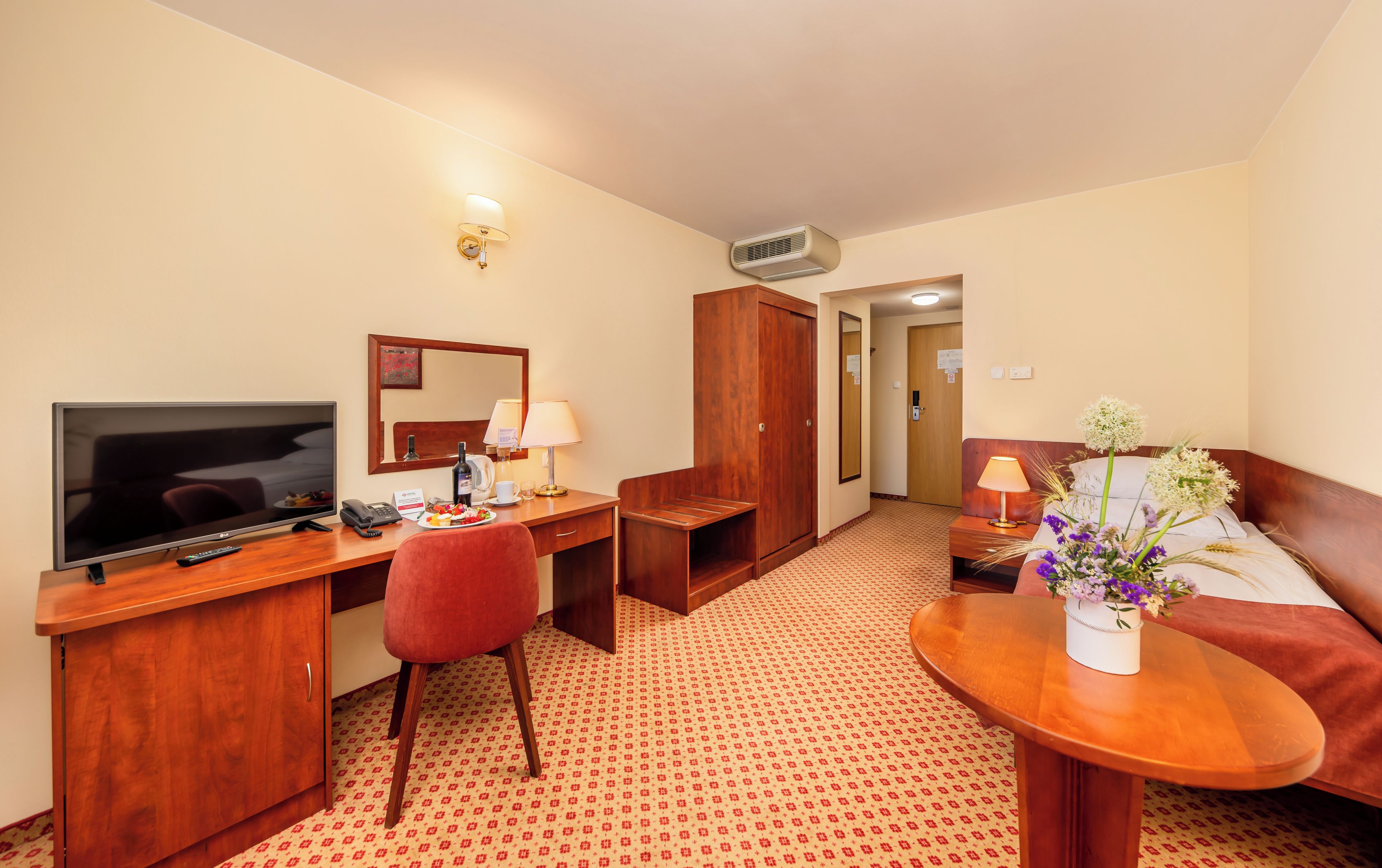 Une chambre d’hôtel équipée d’une télévision, d’un bureau, d’une chaise, d’une armoire, d’une table de chevet, d’une lampe et d’un arrangement floral.