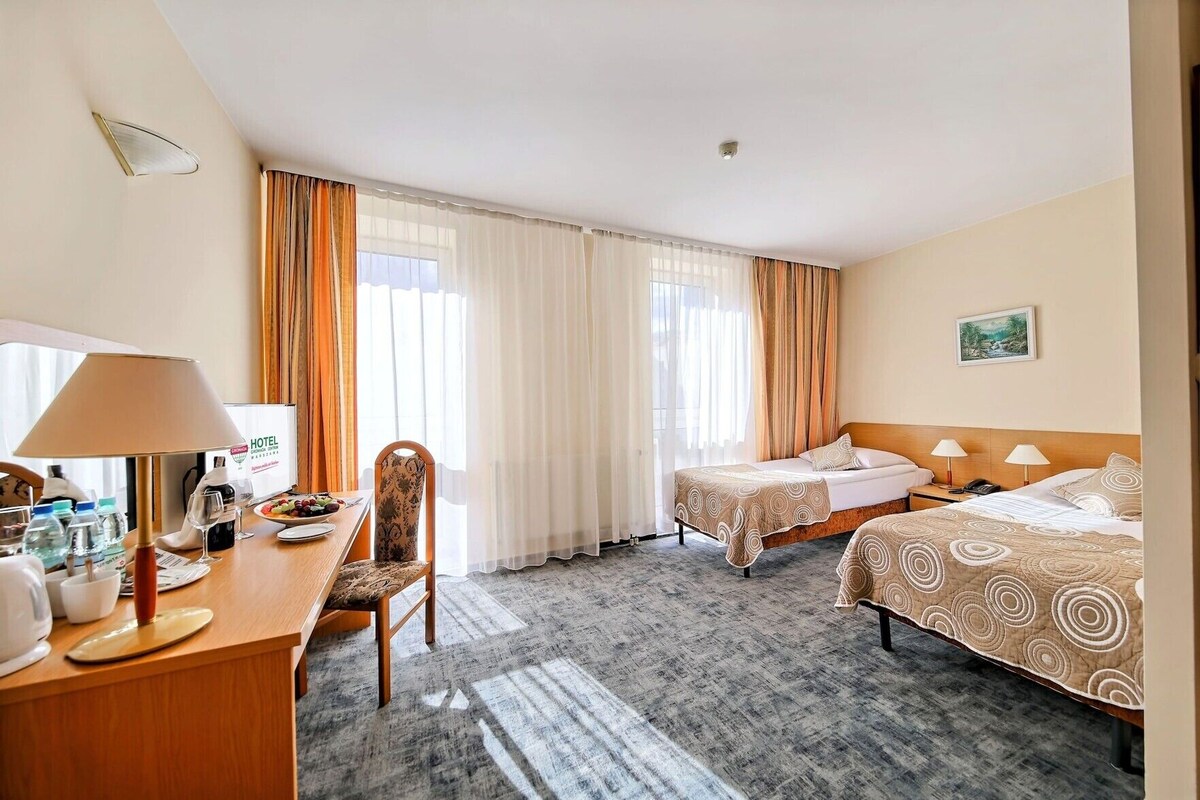 A hotel room with two beds, a desk with a lamp, a chair, and a dining table set with glasses and a bottle.