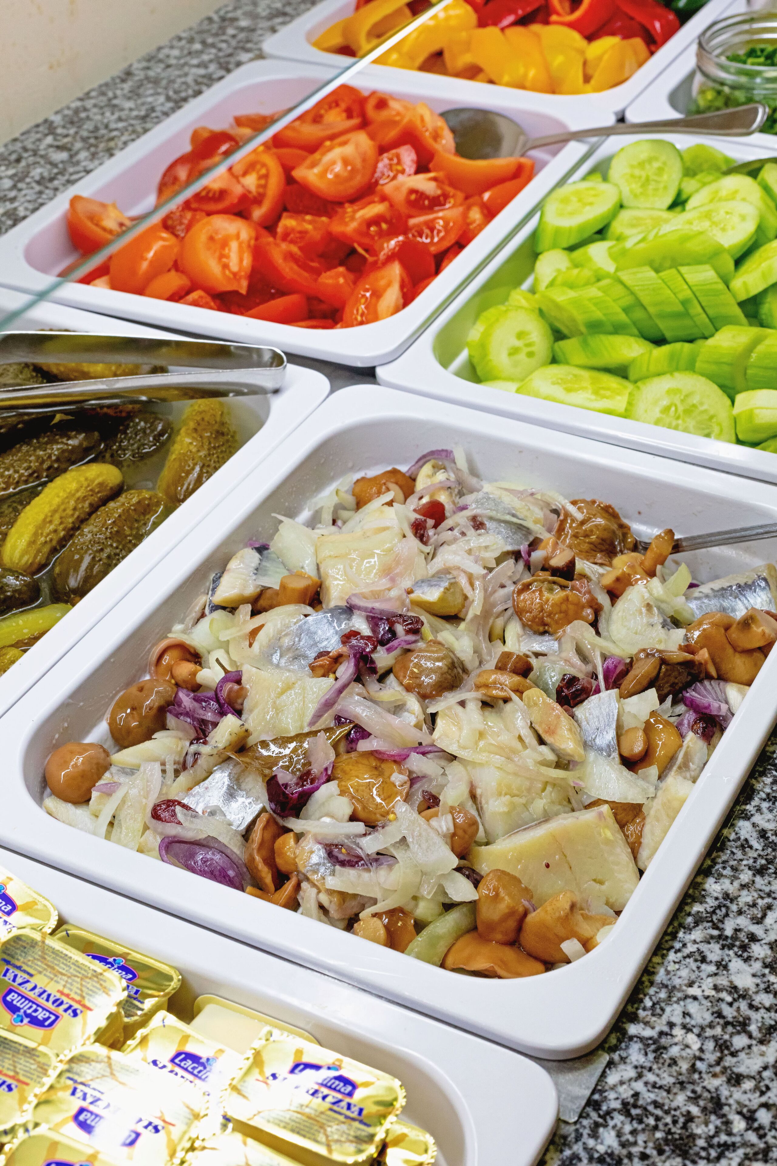 A buffet with various food items including sliced tomatoes, cucumbers, pickles, and a salad with nuts and onions.