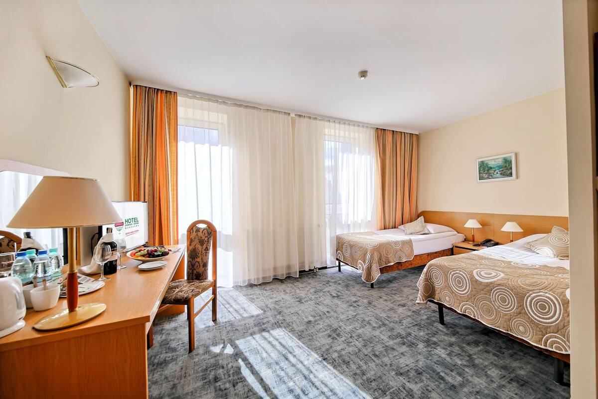 A hotel room with two beds, a desk with a lamp, a chair, and a dining table set with glasses and plates.