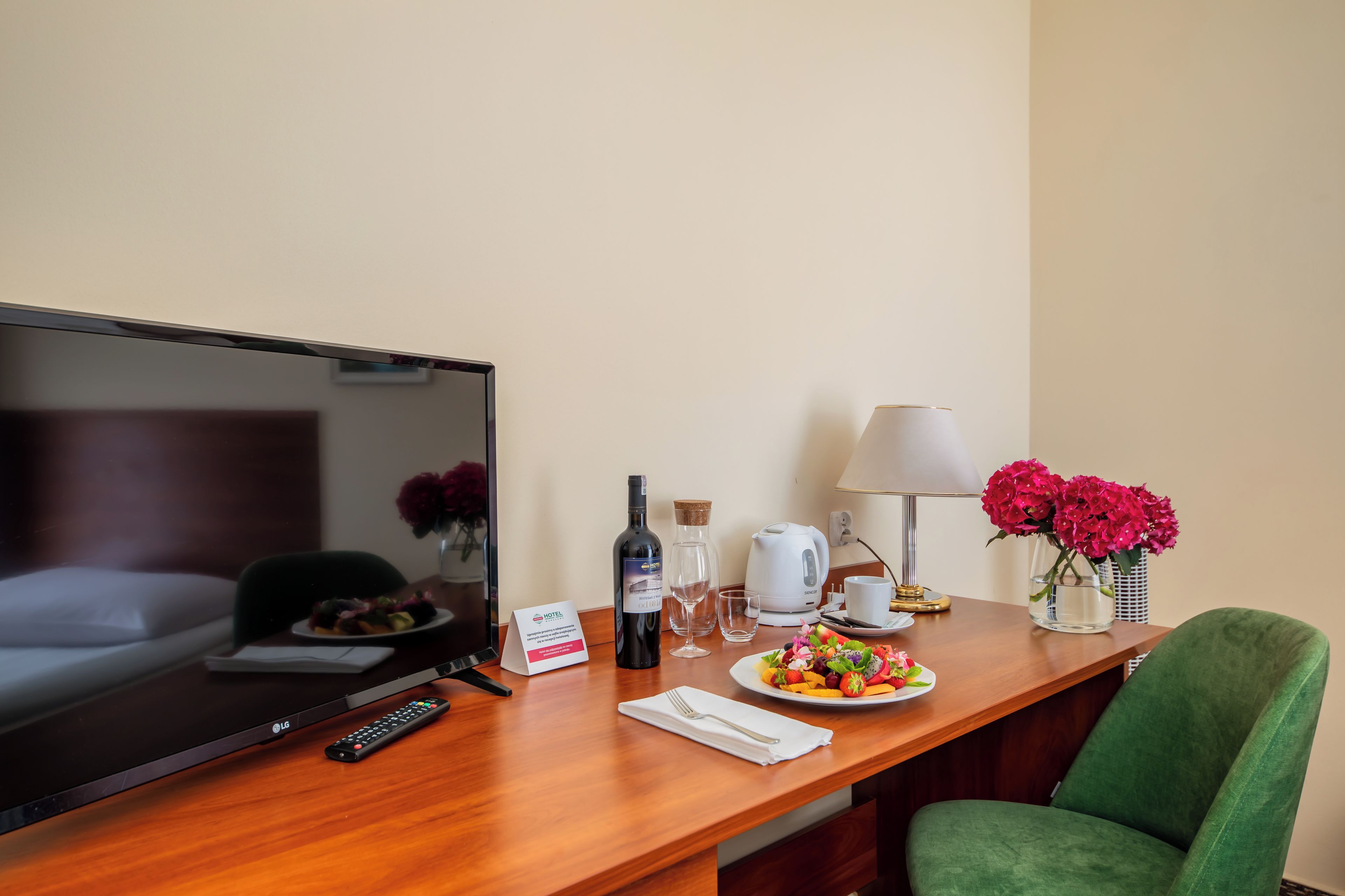 Une chambre d’hôtel avec un bureau en bois, une télévision, une télécommande, une bouteille de vin, une machine à café blanche, un verre d’eau, une assiette de fruits, un vase avec des fleurs et une chaise verte.