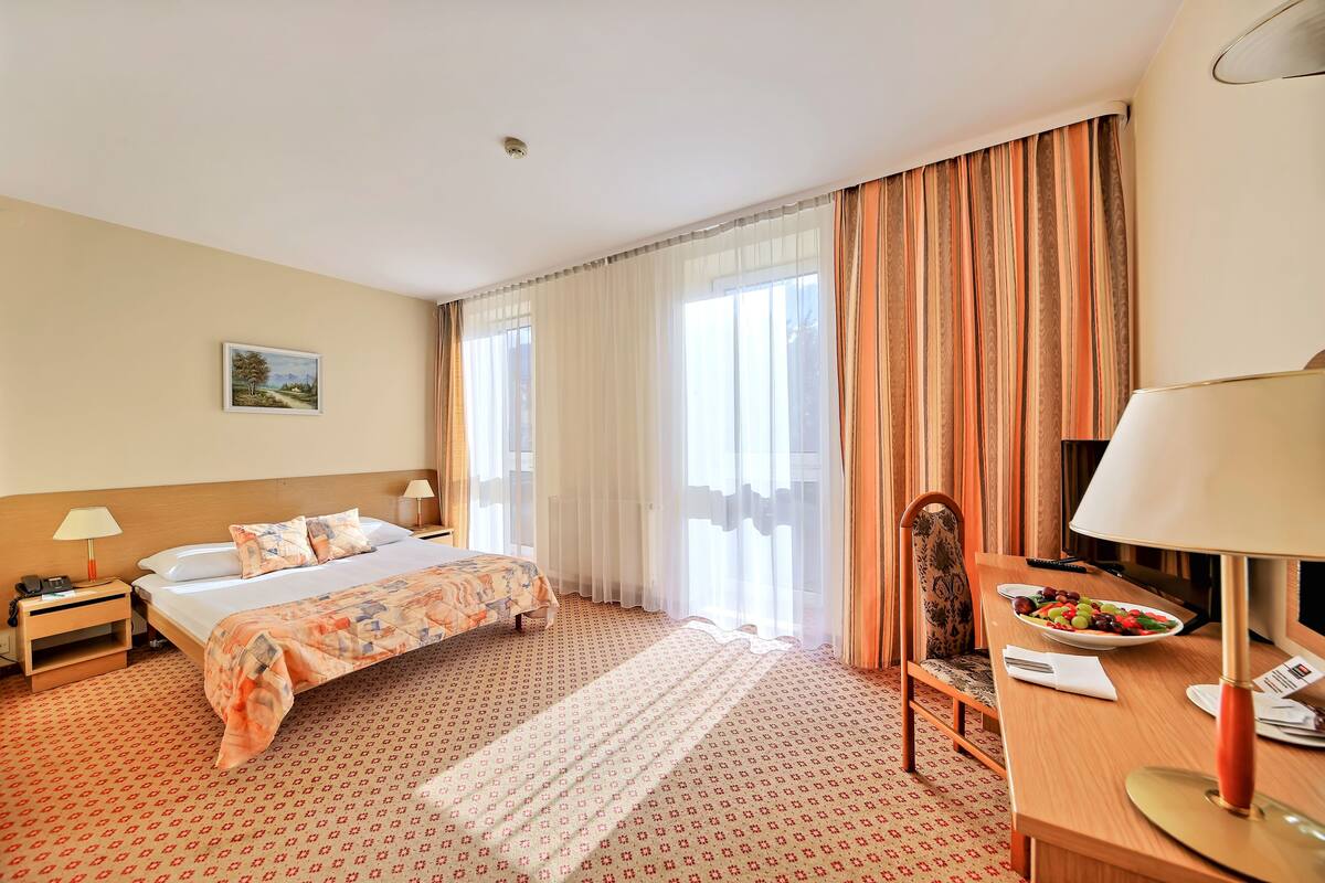 A hotel room with a bed, a desk with a lamp, a chair, a small table with a plate of fruit, and a painting on the wall.