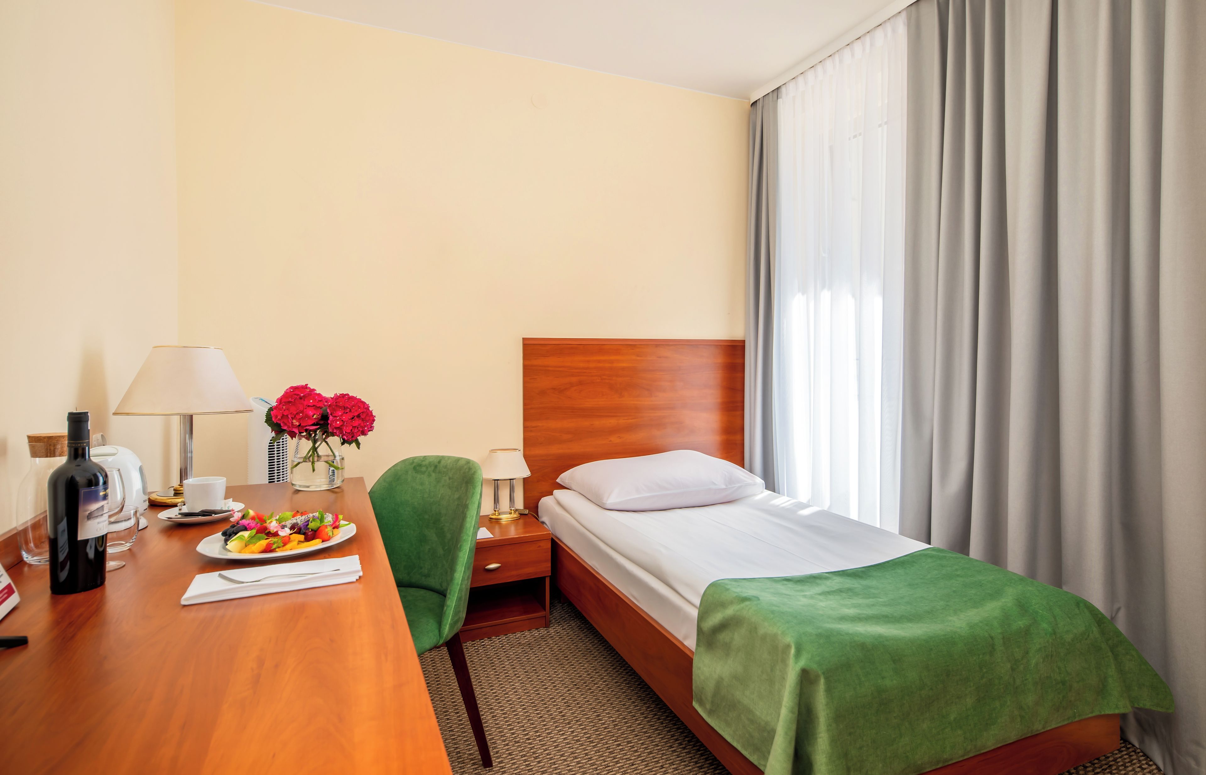 A hotel room with a wooden bed, a green chair, a desk with a plate of fruit, a bottle of wine, and a vase of flowers.